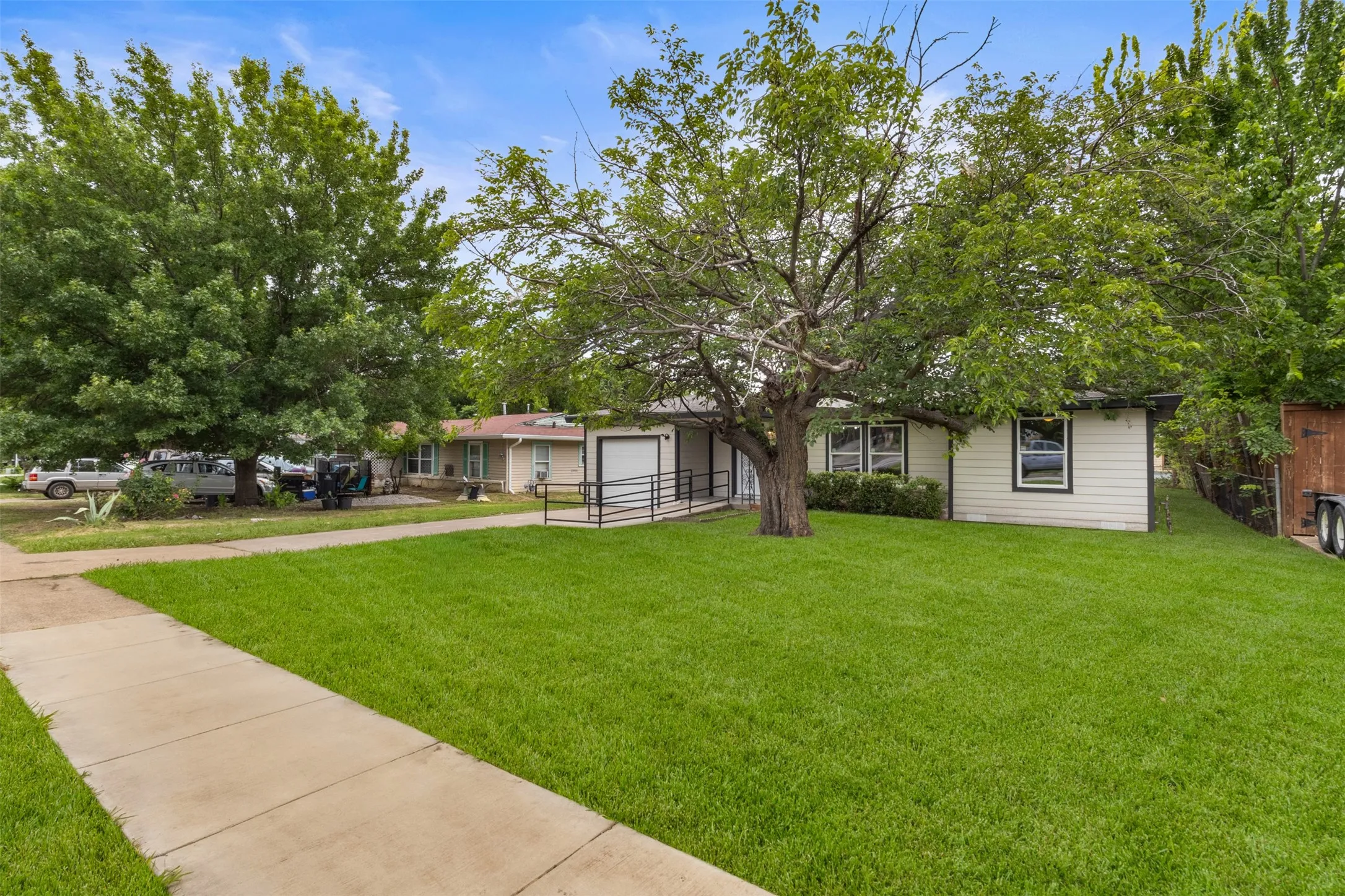 Front exterior showcasing a broad lawn, mature trees, a concrete sidewalk, a ramp entry, and vinyl siding