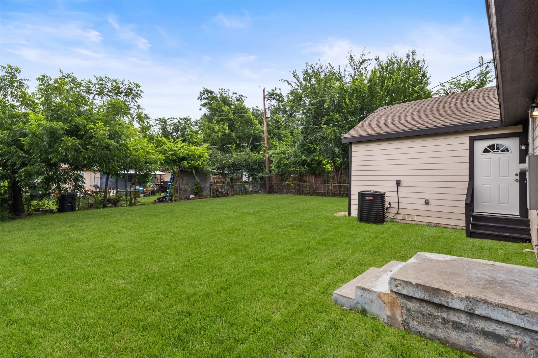 Expansive grassy backyard featuring mature perimeter trees, a concrete patio, and a utility access door