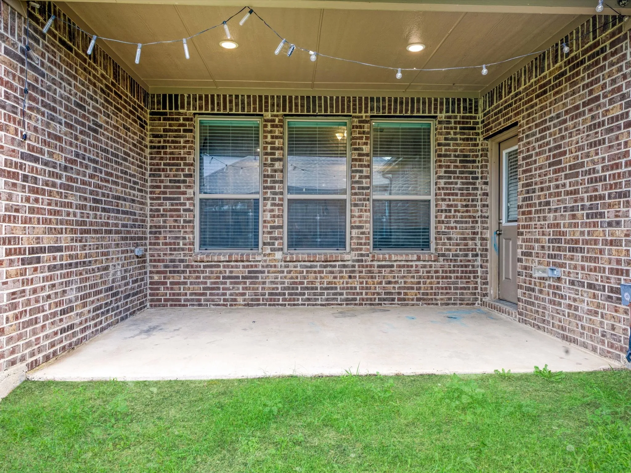 Covered patio featuring brick exterior walls, a concrete slab, three windows with blinds, a side entry door, and recessed ceiling lighting