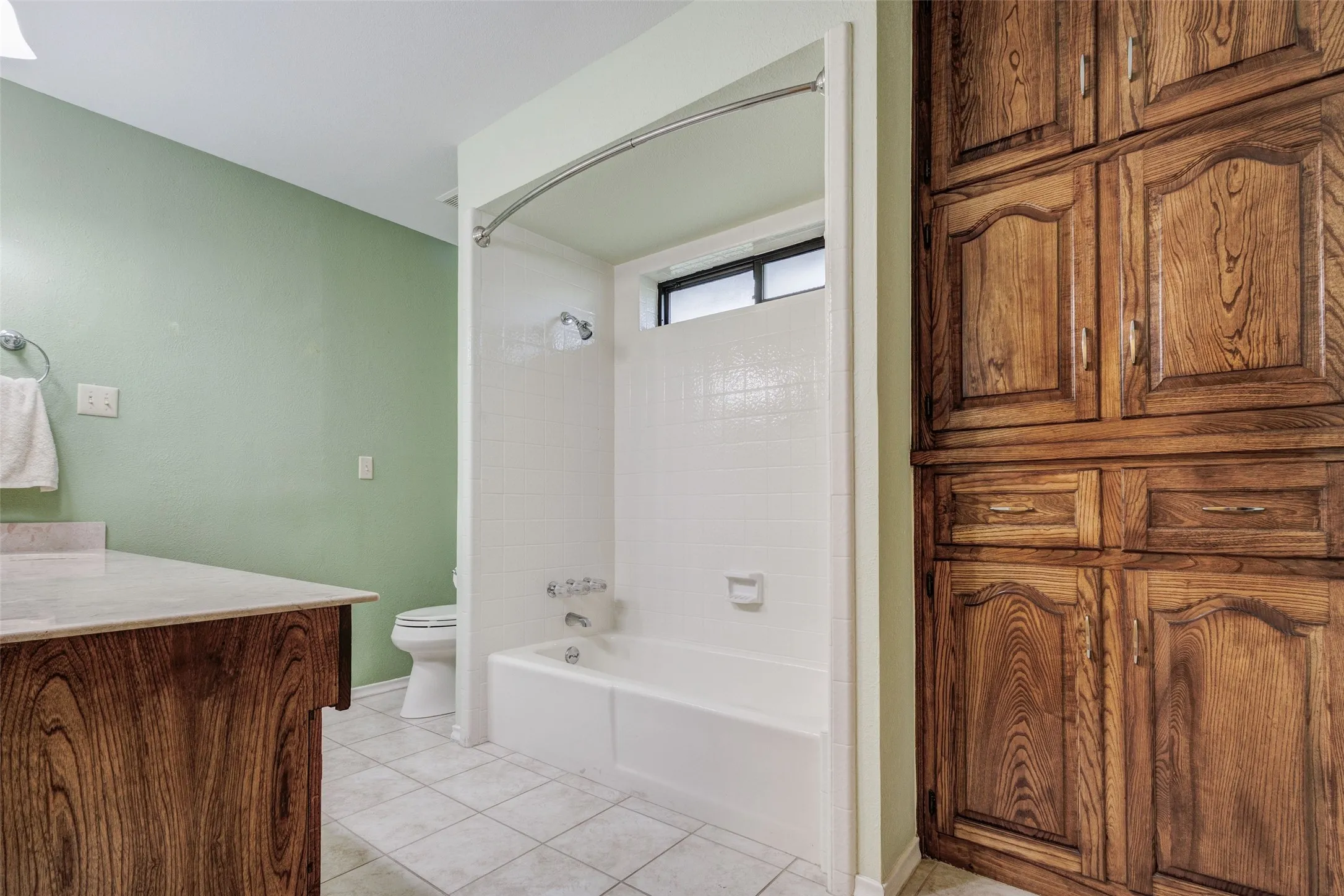 Bathroom featuring a built-in wood-finish vanity with a light-toned countertop, a tub-shower combination with white tile surround, a toilet, and extensive built-in wood-finish cabinetry