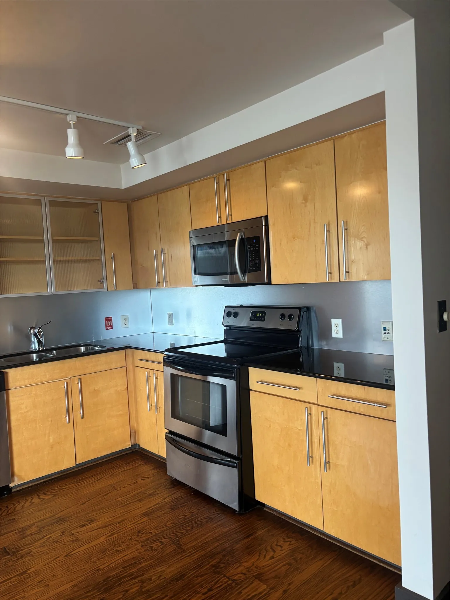 Kitchen featuring light wood cabinetry with brushed nickel hardware, stainless steel appliances, black countertops, and a metallic backsplash