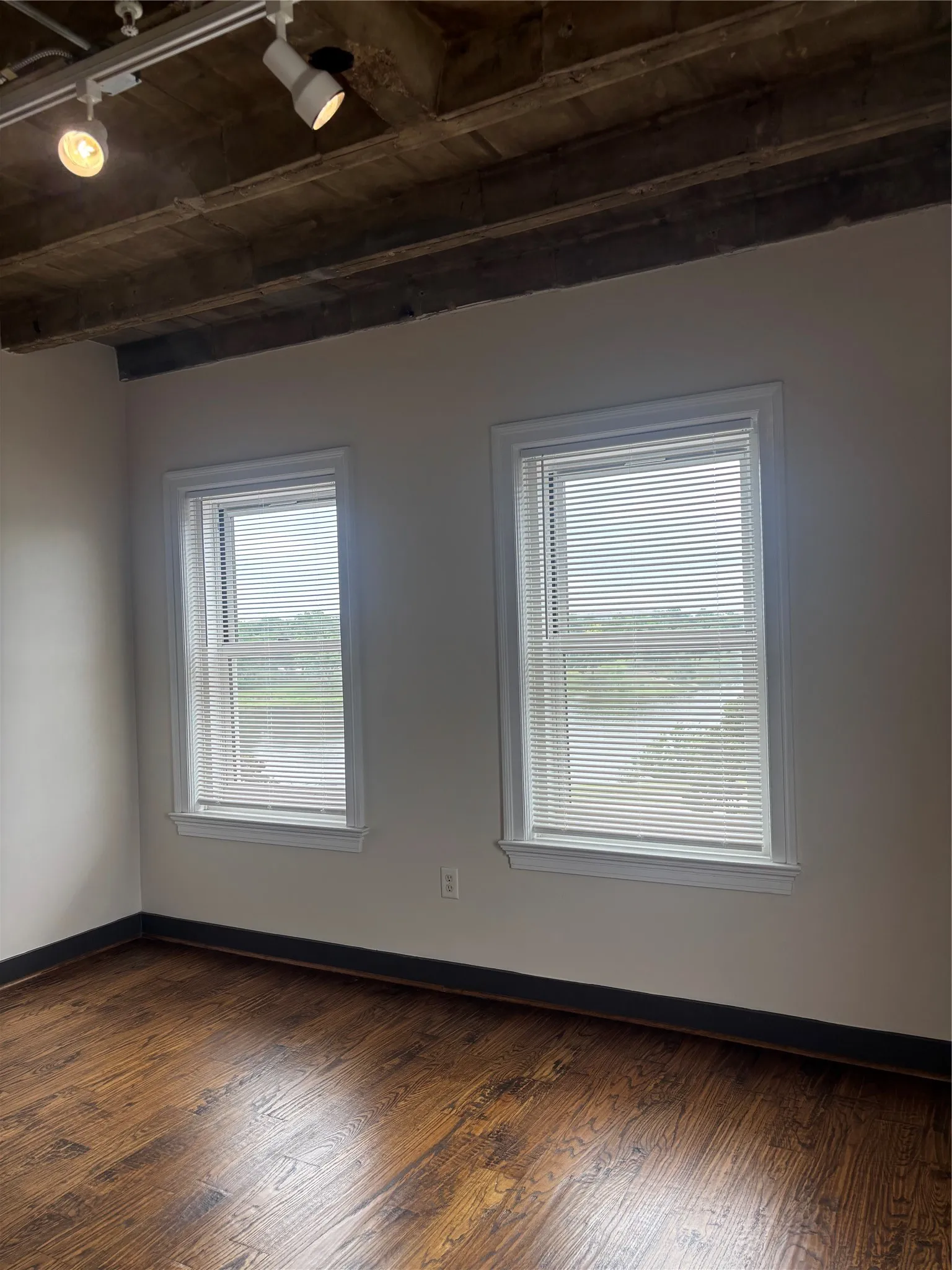 Spacious room featuring wood-finish flooring, two windows with blinds, and white wall paint