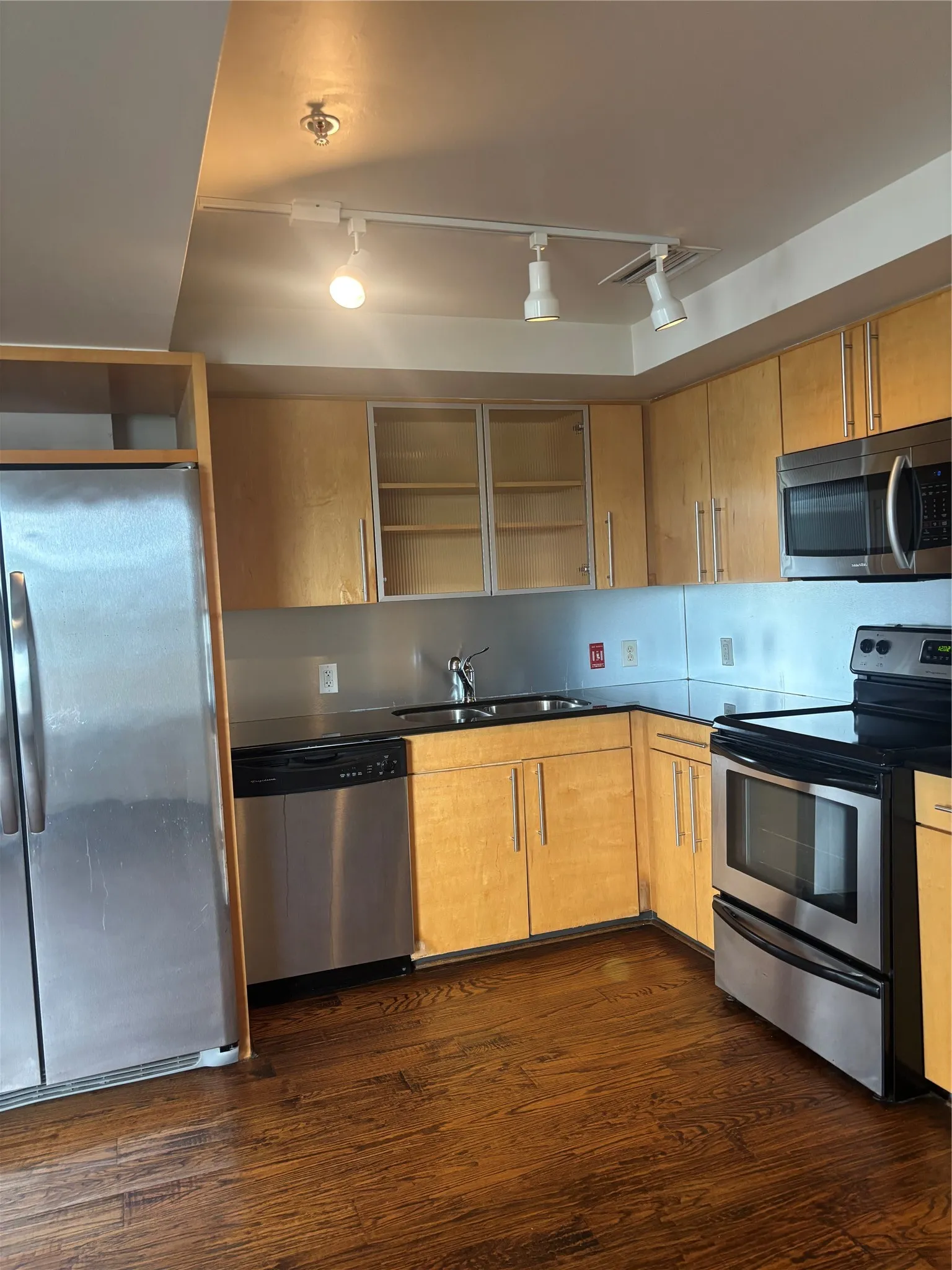 Kitchen featuring wood-finish flooring, stainless steel appliances, black countertops, track lighting, and light wood cabinetry
