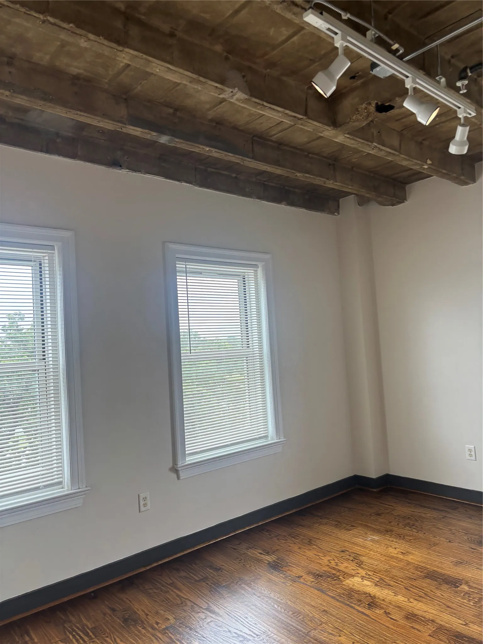 Space featuring two windows with white blinds, track lighting, exposed concrete ceiling, and wood-finish flooring