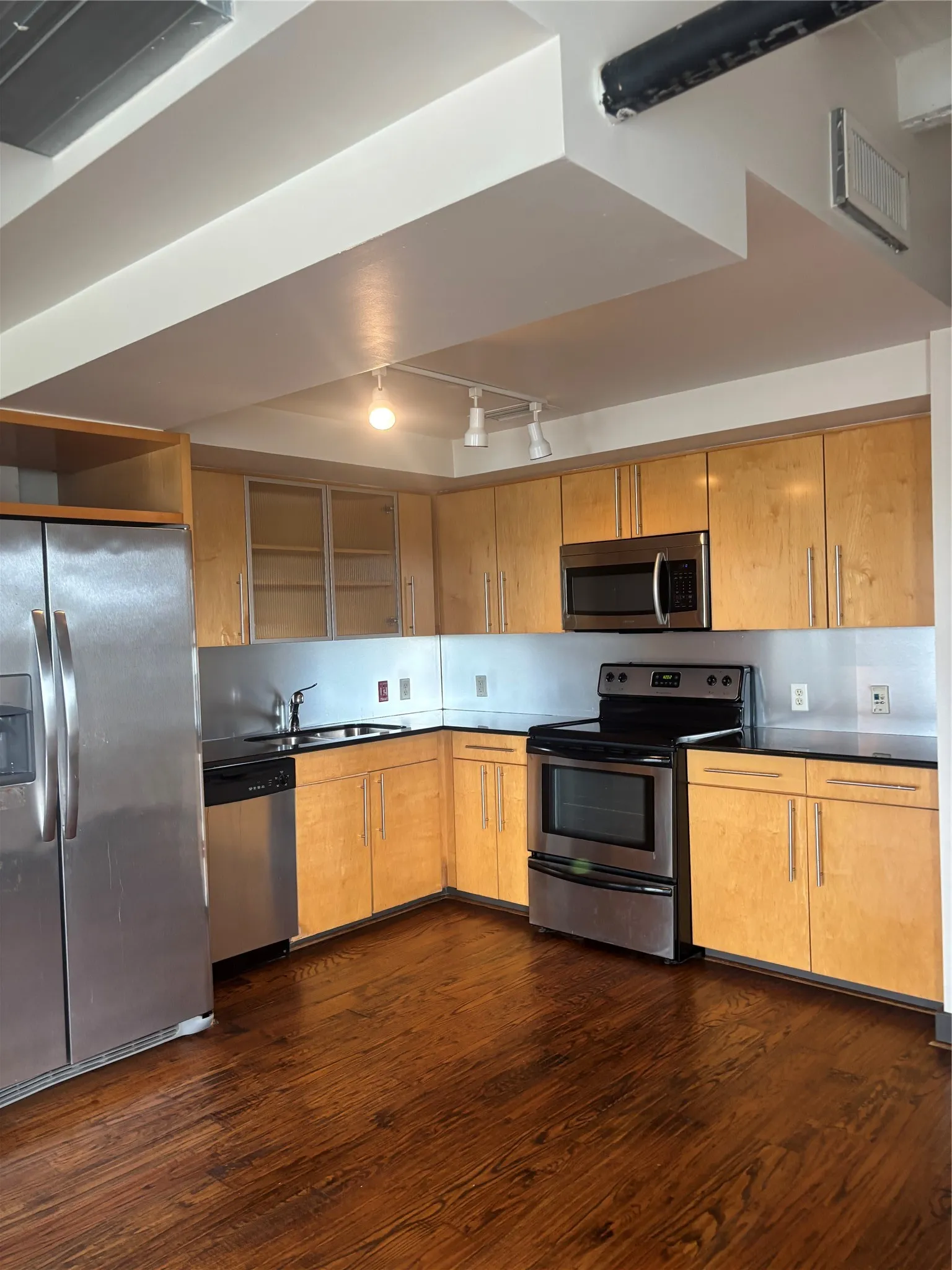 Modern kitchen featuring stainless steel appliances, wood-finish flooring, light wood cabinetry, dark countertops, and exposed ductwork