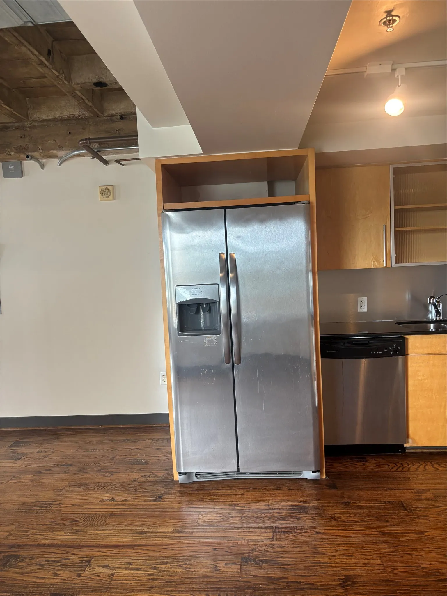 Kitchen area featuring wood-finish flooring, stainless steel side-by-side refrigerator, stainless steel dishwasher, light wood cabinetry, and dark countertops