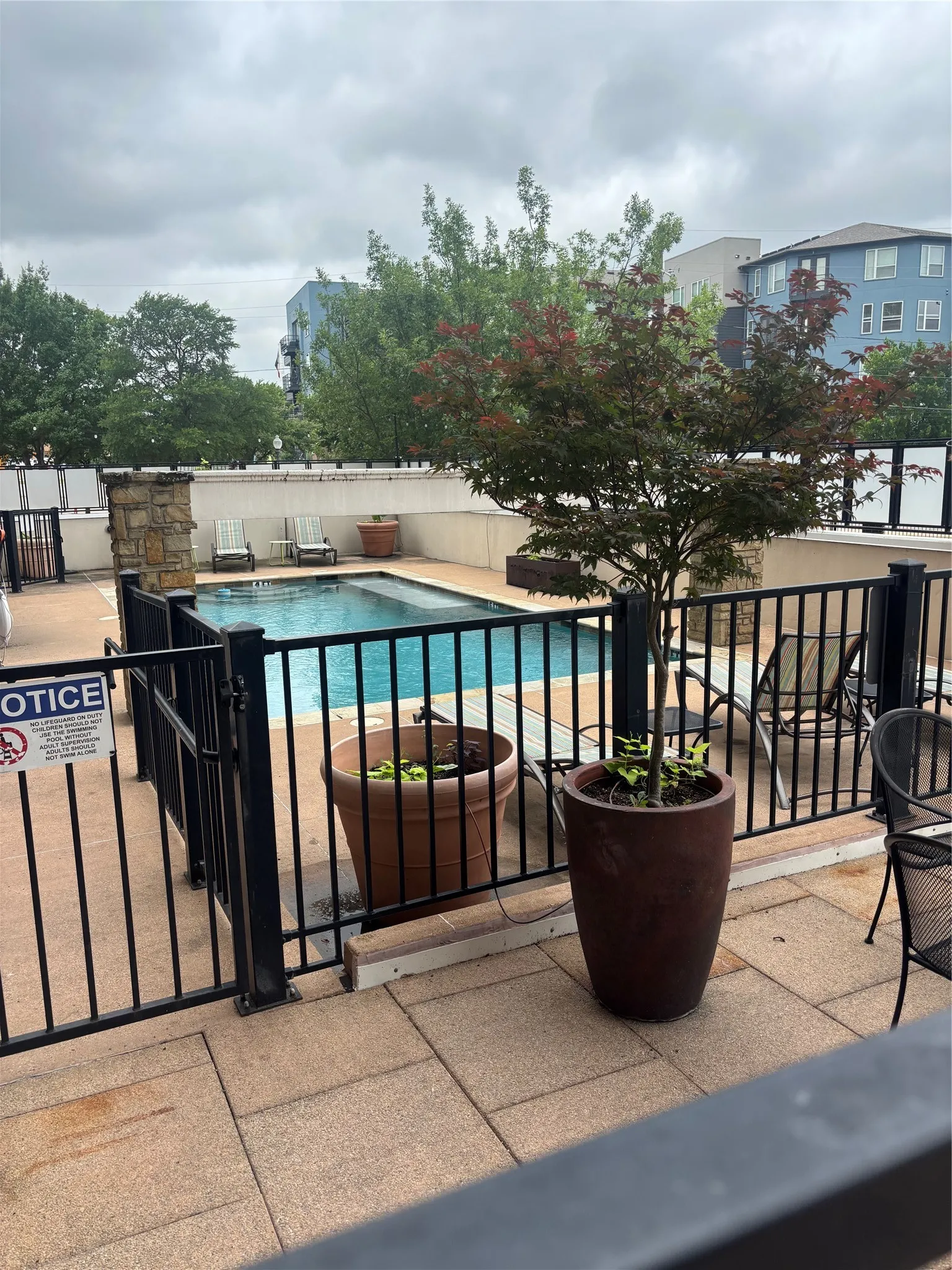 Resort-style pool area featuring a perimeter fence, stone-clad pillars, and a tiled sundeck with potted plants