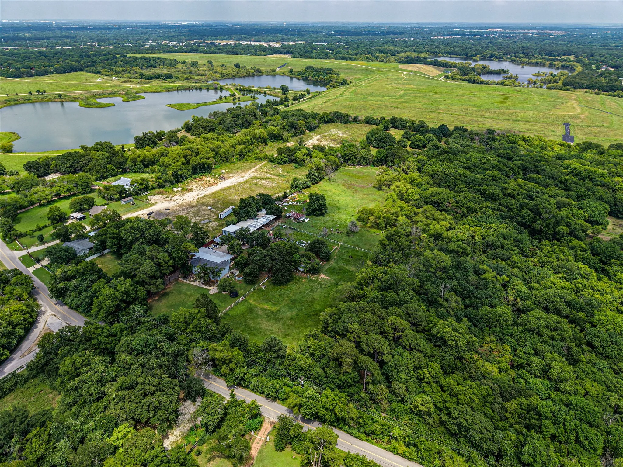 Expansive aerial perspective showcasing significant land acreage with multiple structures, mature tree cover, and a paved road frontage