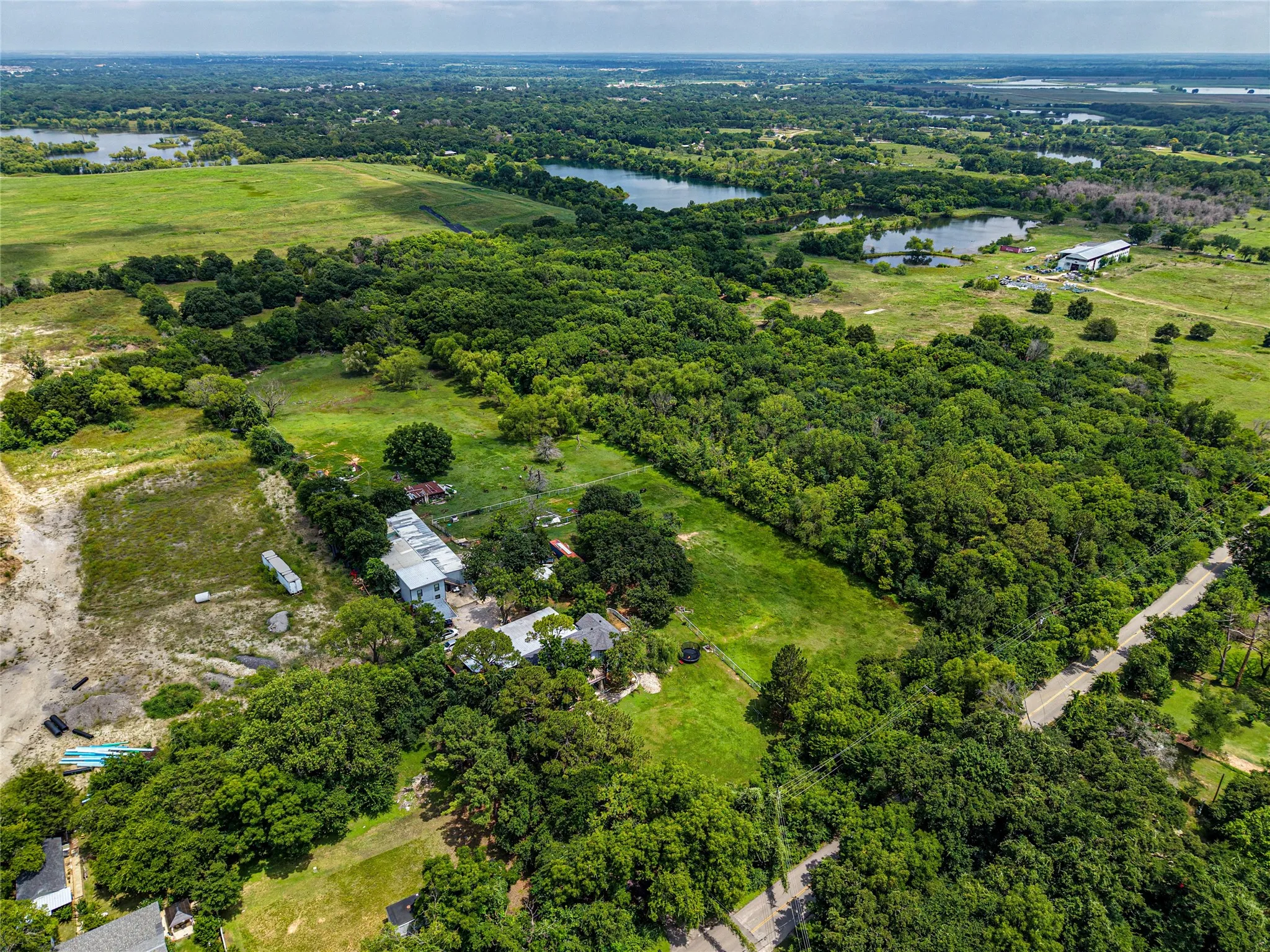 Expansive rural property featuring extensive tree coverage, open grassy areas, multiple bodies of water, and a paved road bordering the property