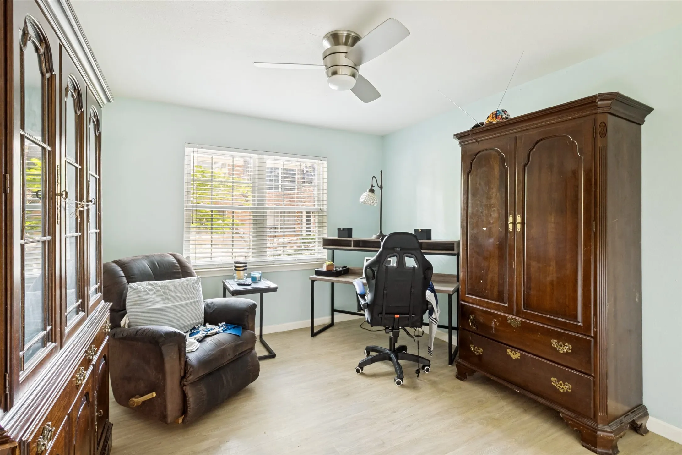 Light blue walls complement the light-toned wood-finish flooring in this room featuring a window with blinds, a ceiling fan, and ample space for furnishings