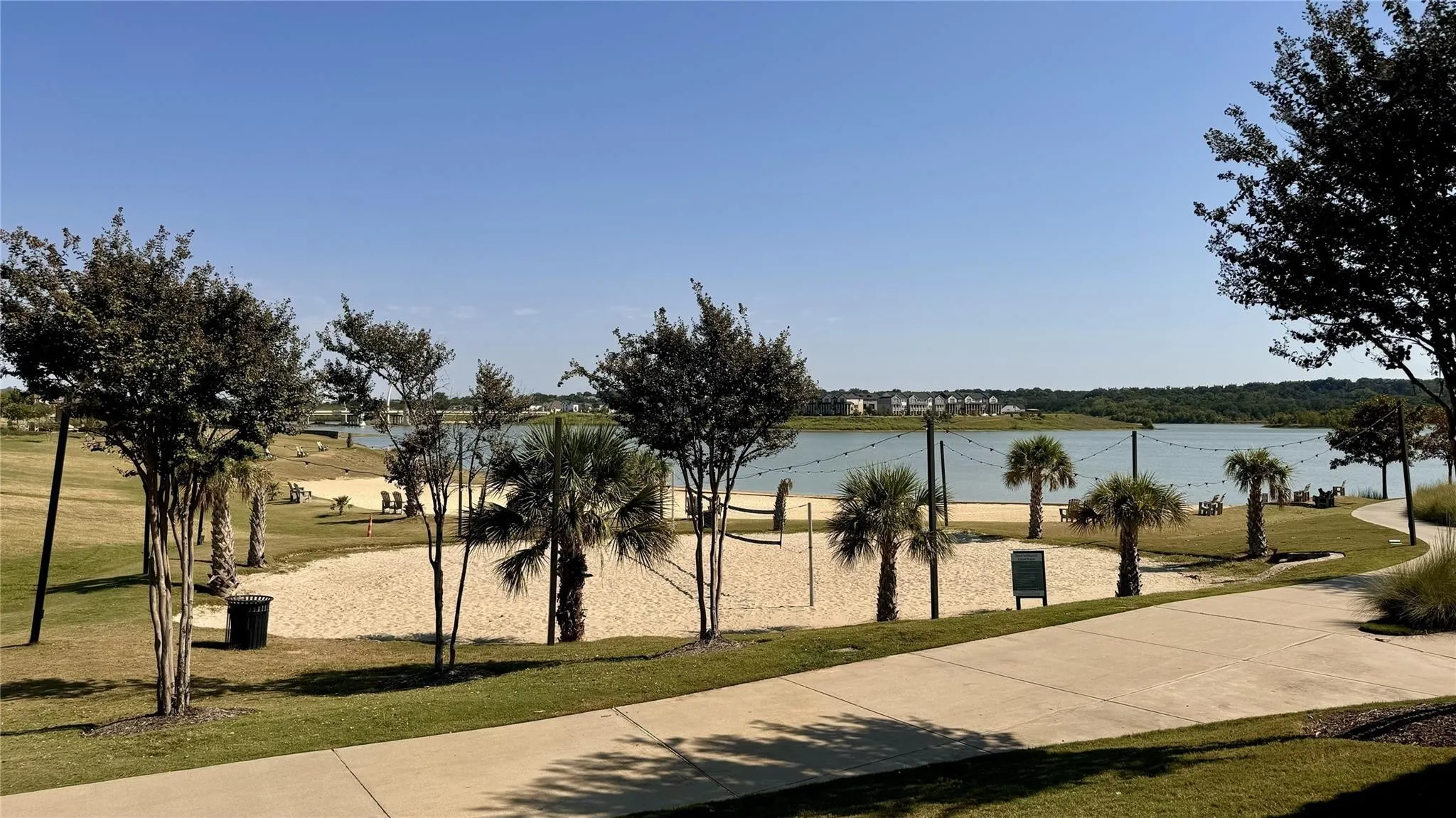 Lakeside recreational area featuring a sand volleyball court, mature palm trees, string lighting, a concrete pathway, and a grassy expanse