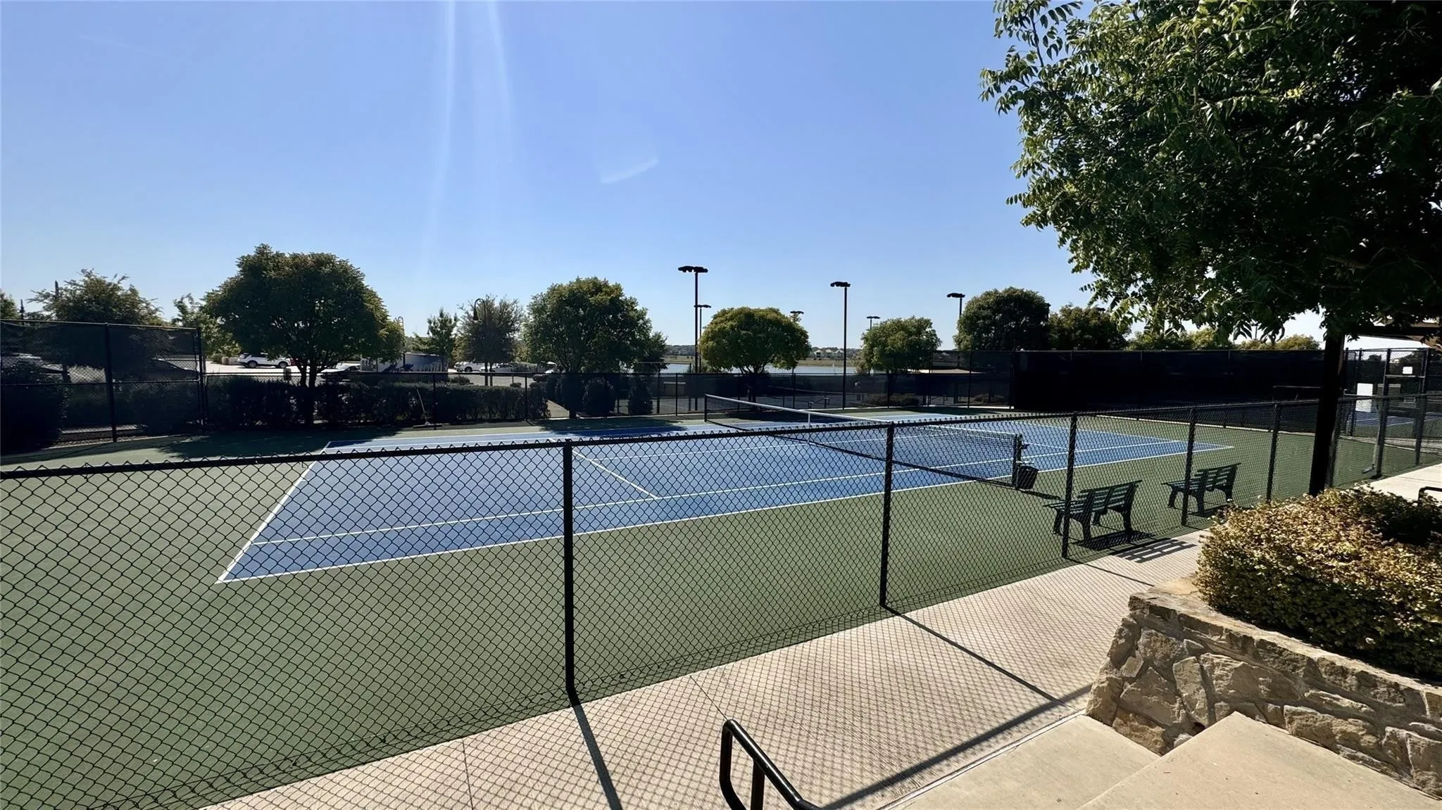 Fenced tennis courts featuring blue and green playing surfaces, court lighting, and surrounding landscaping