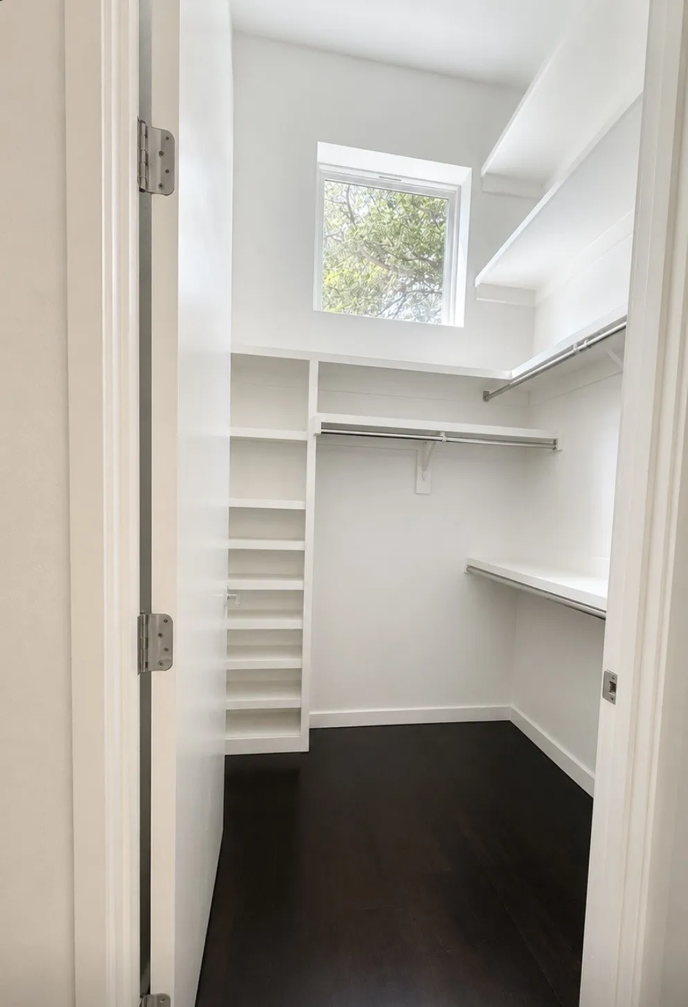 Walk-in closet with dark wood-finish flooring, built-in shelving, hanging rods, and a window for natural illumination