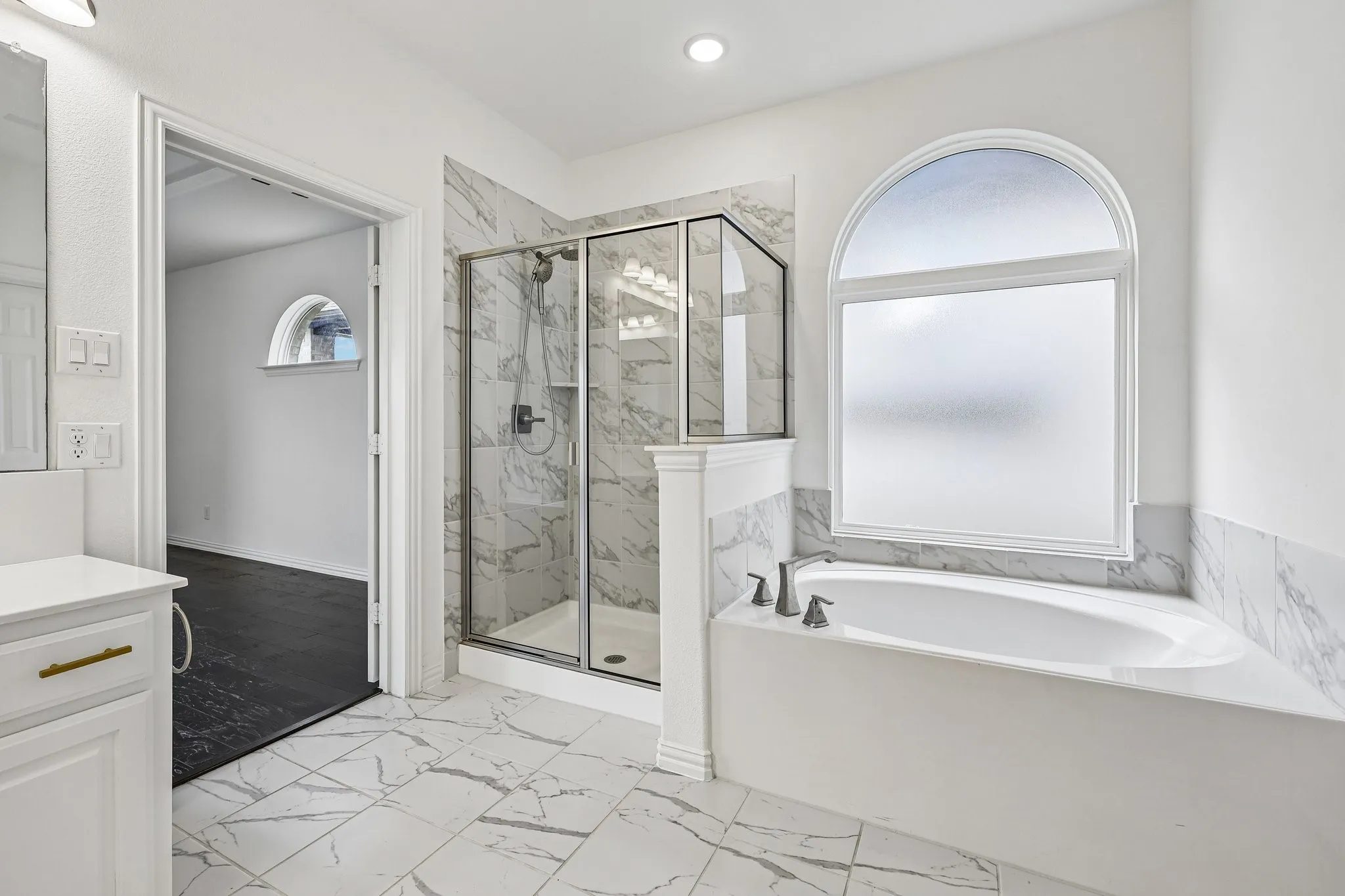 Bathroom featuring a soaking tub with an arched window, a glass-enclosed shower, white vanity with gold-tone hardware, and marble-look tile flooring