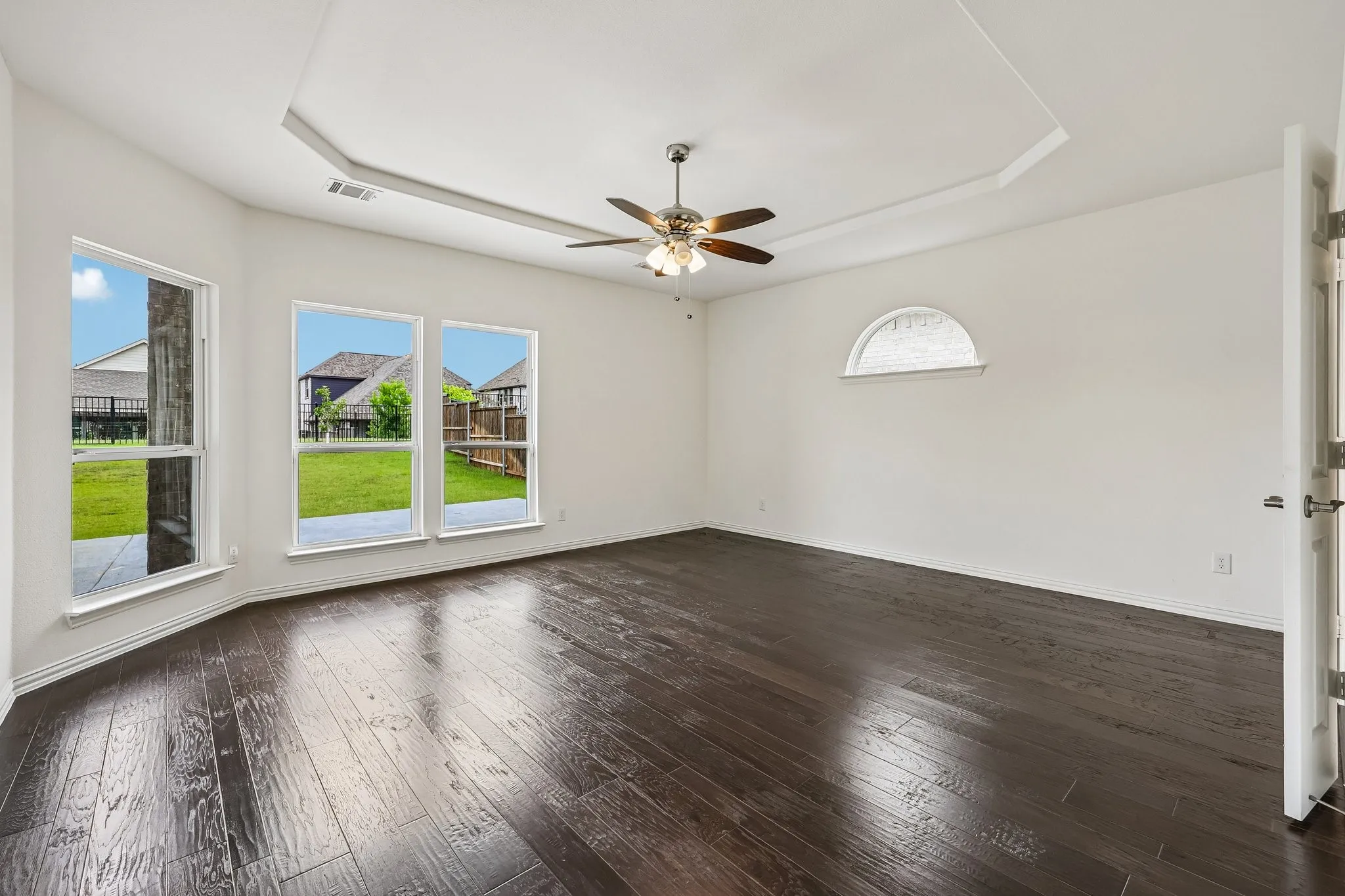 Spacious room featuring dark wood-finish flooring, a tray ceiling with a ceiling fan, multiple windows including an arched window, and a white interior door with contemporary hardware
