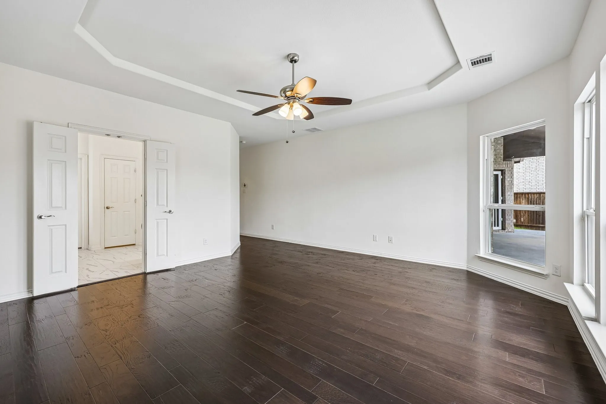 Spacious room featuring dark wood-finish flooring, a tray ceiling with a ceiling fan, and white walls