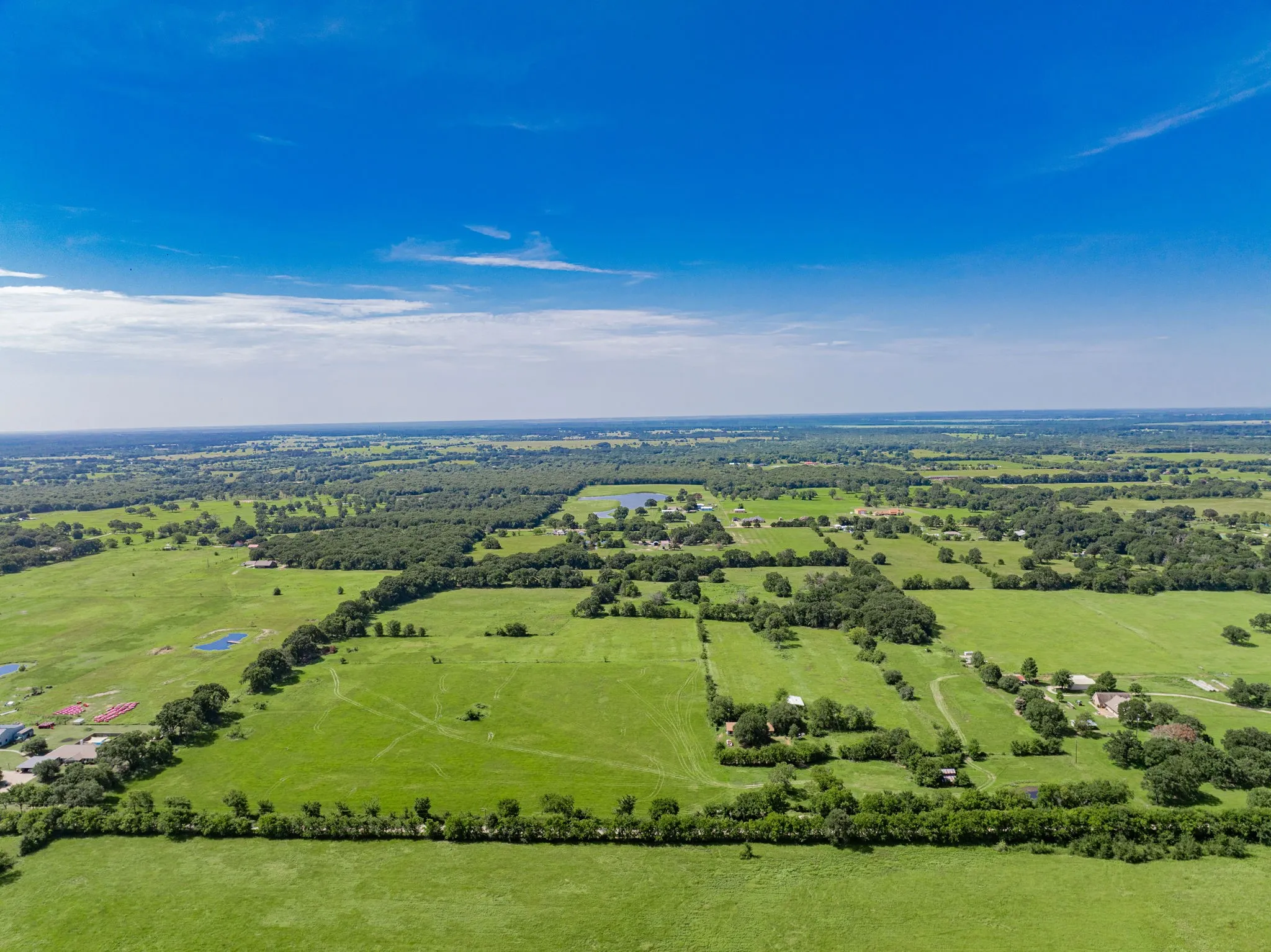 Expansive rural landscape featuring extensive green pastures, scattered mature trees, multiple ponds, and distant residential structures