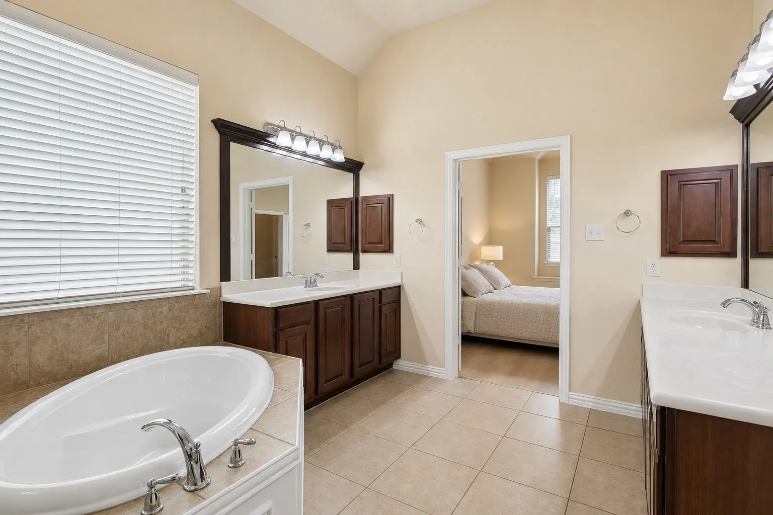 Bathroom featuring a built-in soaking tub with chrome fixtures, dual dark wood vanities with integrated sinks and white countertops, large vanity mirrors, and tile flooring