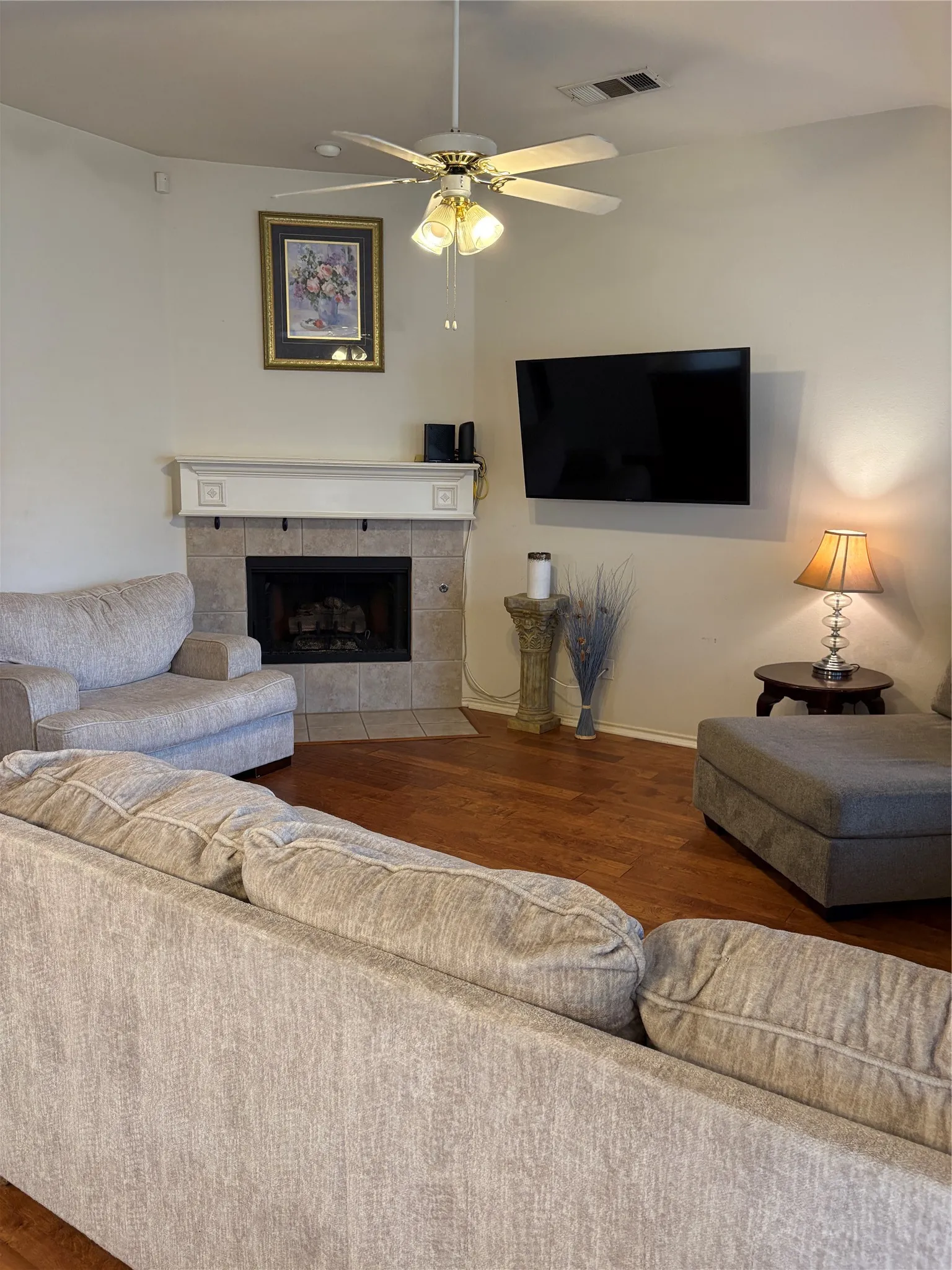 Hardwood flooring, tiled fireplace surround, integrated fireplace, wall-mounted television, and a ceiling fan with light fixture