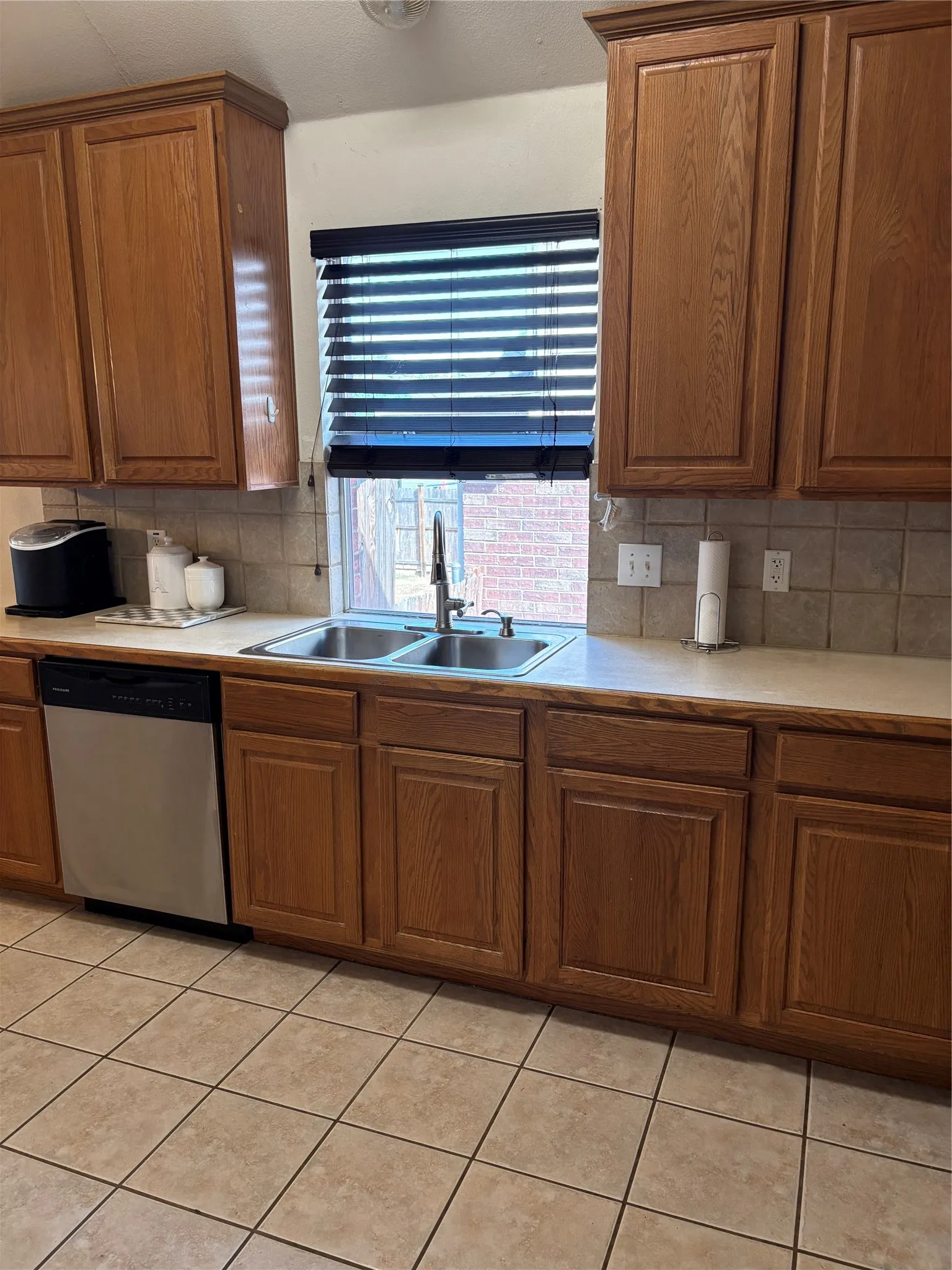Warm wood cabinetry, dual basin stainless steel sink, stainless steel dishwasher, and neutral tile flooring