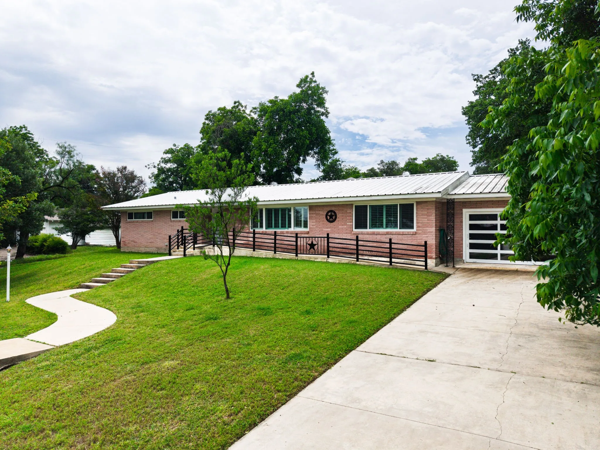 Brick exterior with a white metal roof, featuring a modern garage door with frosted glass panels, a concrete driveway, and a winding sidewalk leading to the entrance