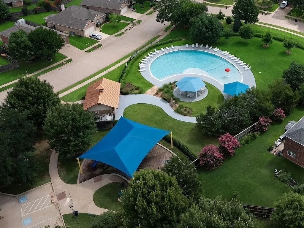 Community amenities featuring an expansive swimming pool, shaded seating areas, a playground with a blue shade structure, and paved walking paths surrounded by mature landscaping