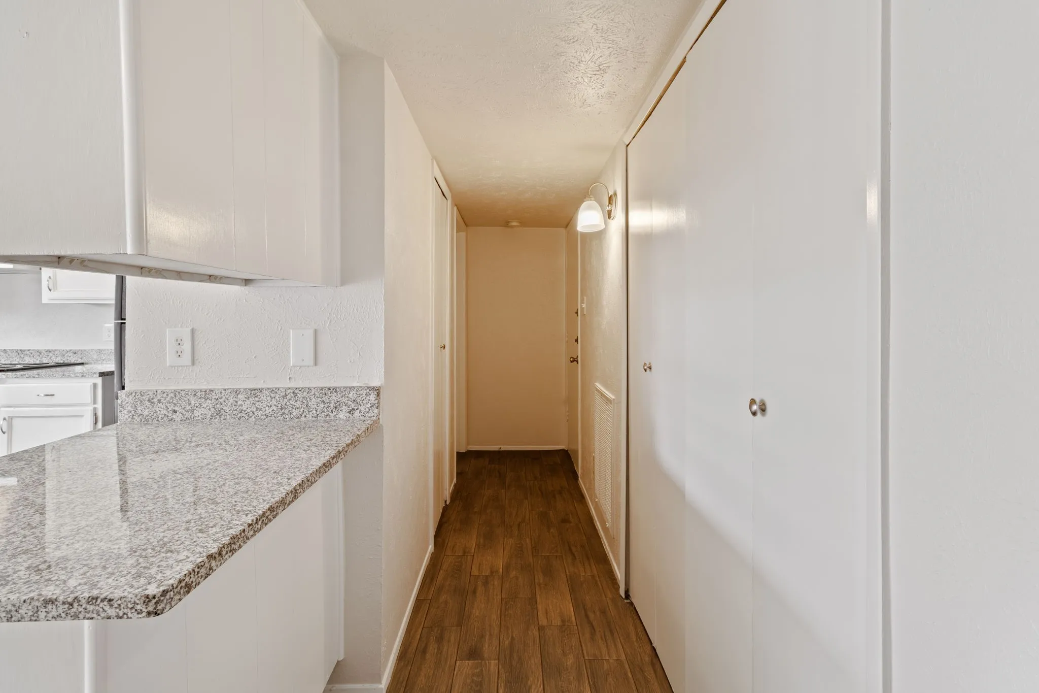 Kitchen peninsula with granite-finish countertop, white cabinetry, wood-finish flooring, and hallway access