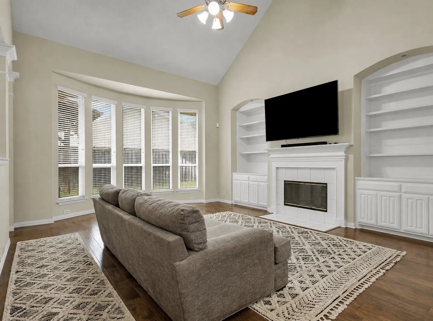 Vaulted ceiling with a ceiling fan, bay window, tile fireplace with a mantel, built-in shelving, and wood-finish flooring