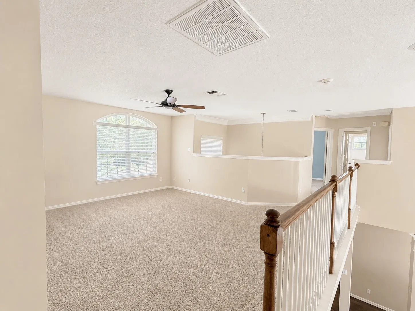 Expansive carpeted loft space featuring an arched window, ceiling fan, and painted baluster railing