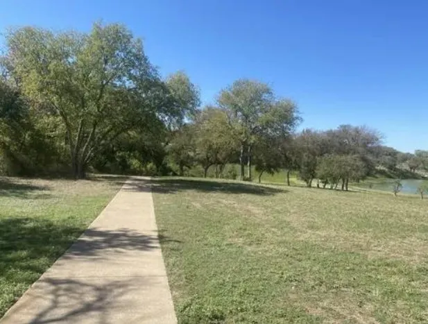 Paved pathway through a grassy expanse with mature trees and water feature