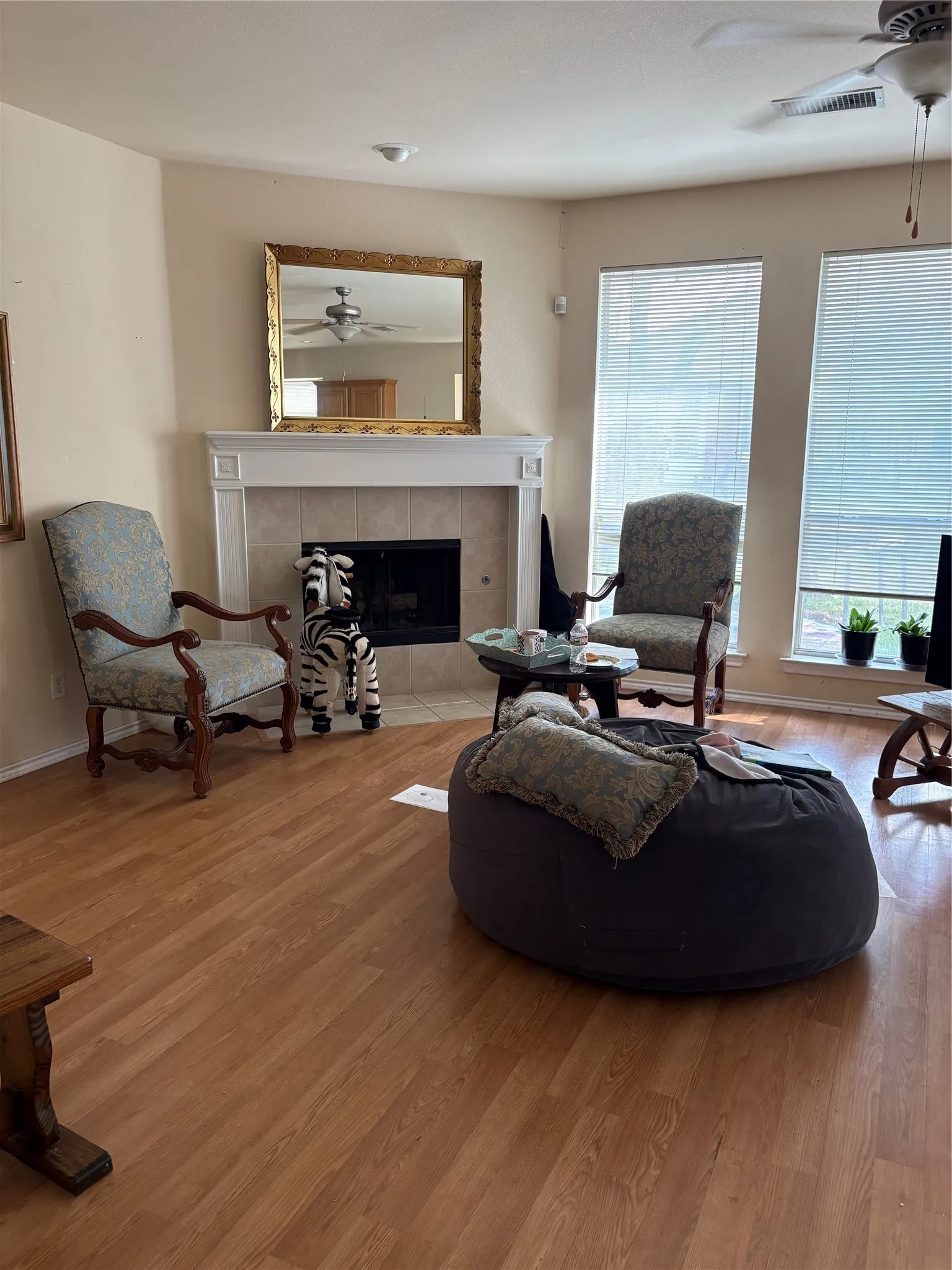 Wood-finish flooring, fireplace with a tile surround and white mantel, ceiling fan, and multiple windows with blinds