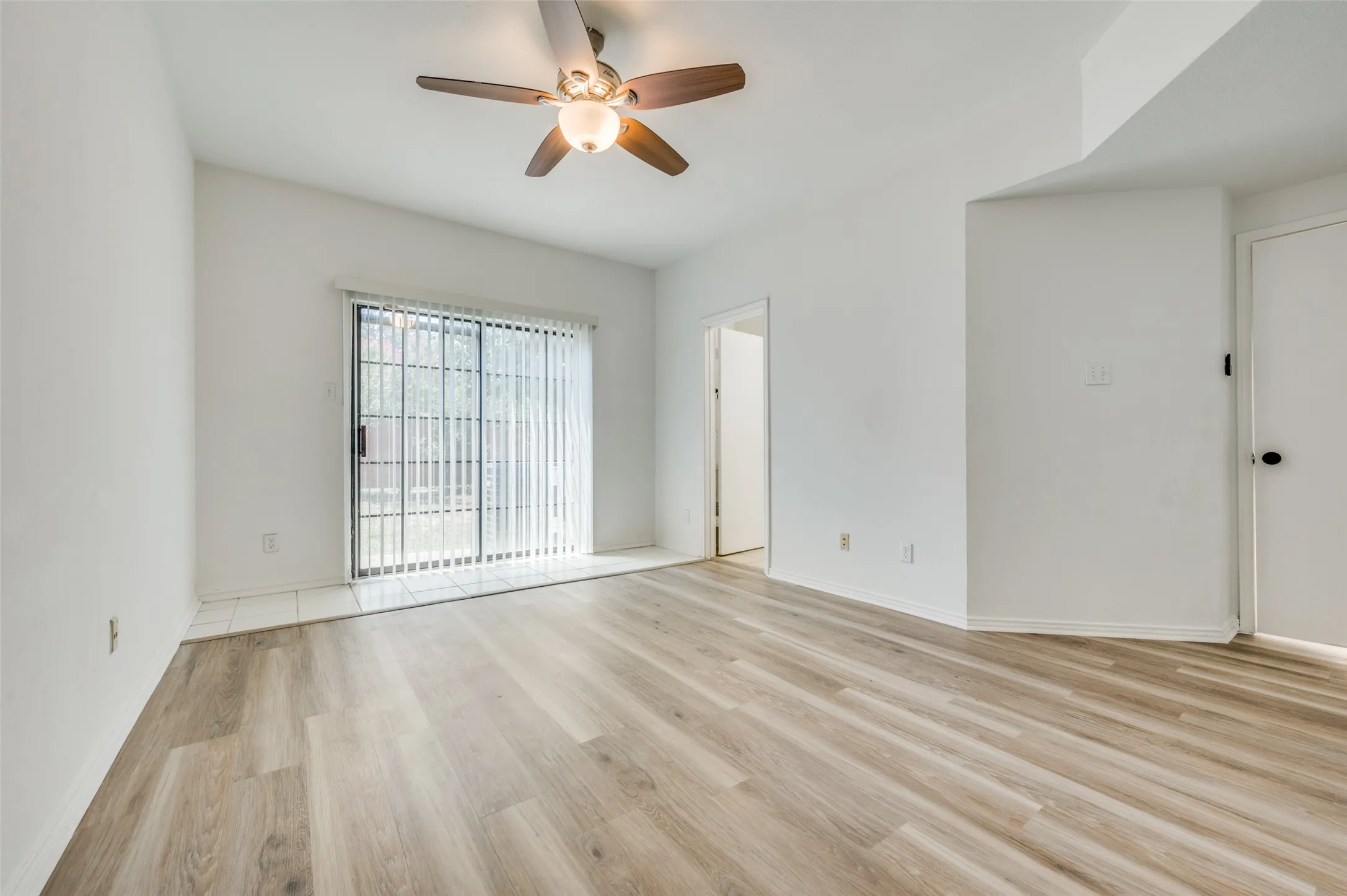 Spacious room featuring wood-finish flooring, a ceiling fan with integrated lighting, and a sliding glass door with vertical blinds