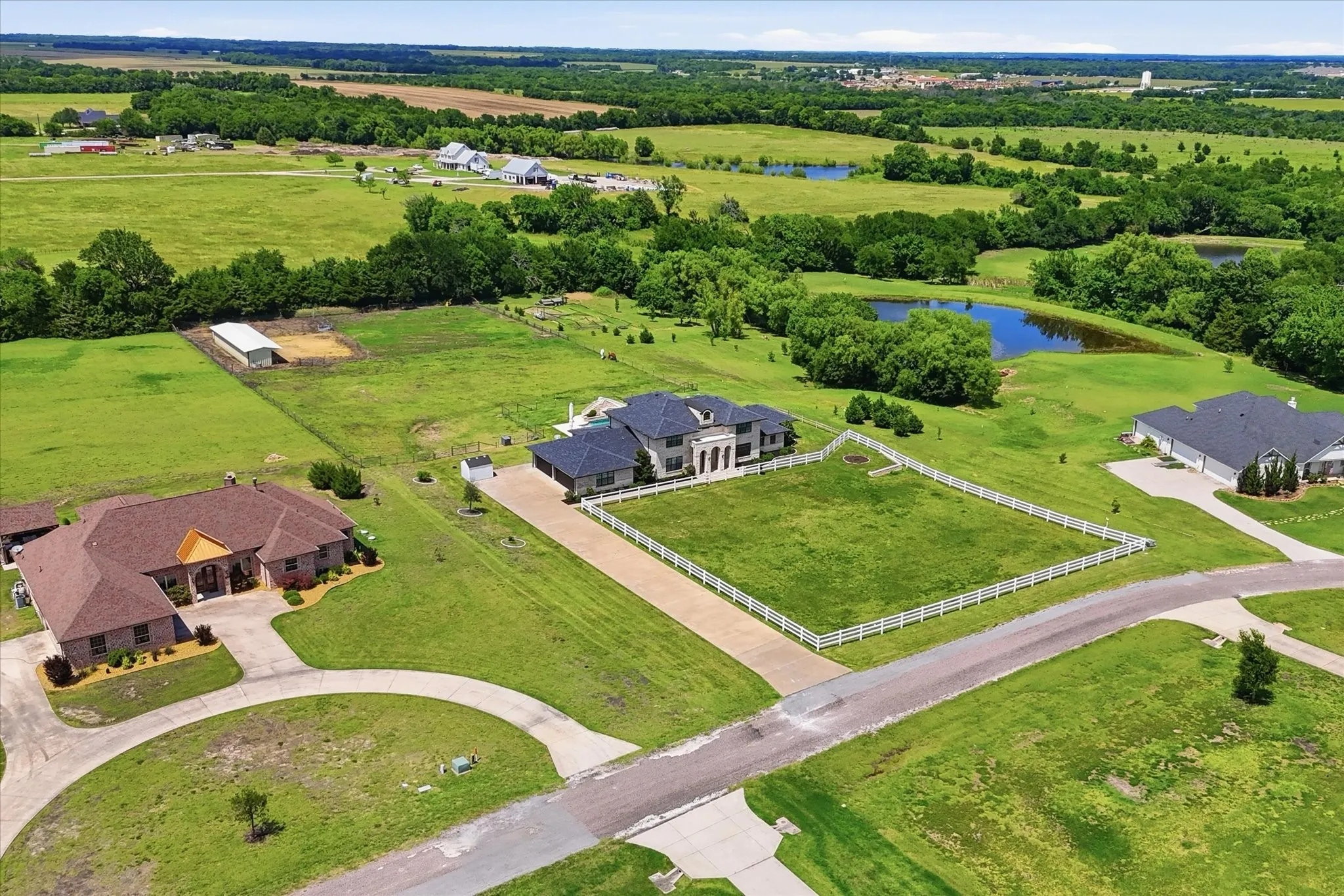 Spacious property featuring a large residence with an expansive driveway, a white perimeter fence, mature trees, and a pond