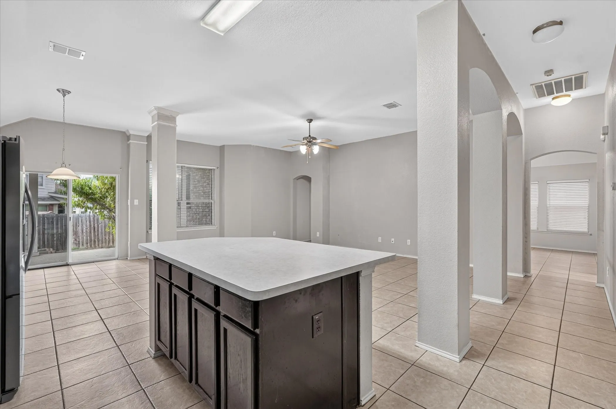Spacious open-concept living area featuring a kitchen island with storage, tile flooring throughout, multiple arched doorways, a ceiling fan, and abundant natural light from a sliding glass door and windows