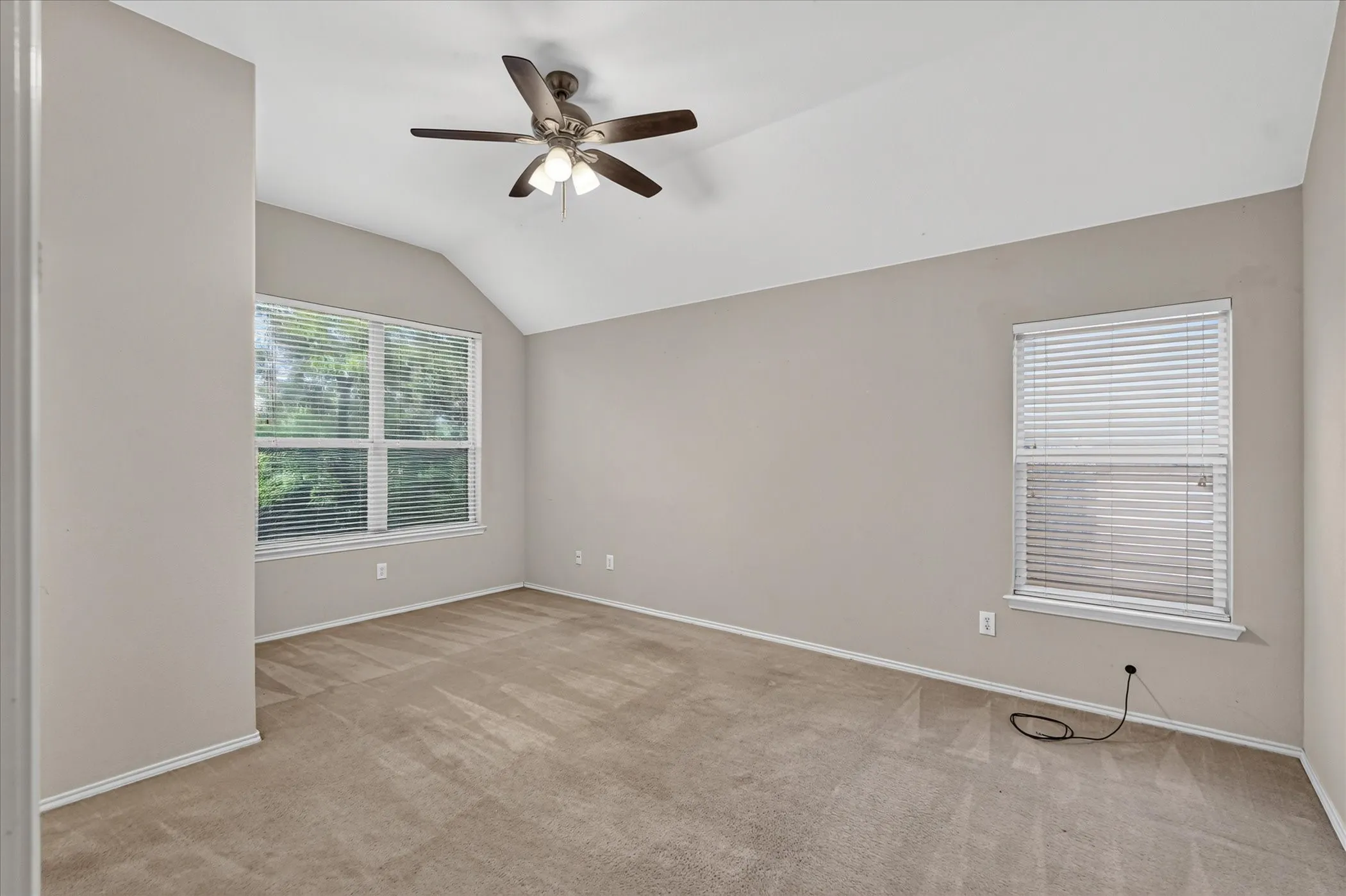 Spacious room featuring vaulted ceilings, a ceiling fan with integrated lighting, two windows with blinds, and neutral-toned carpeting