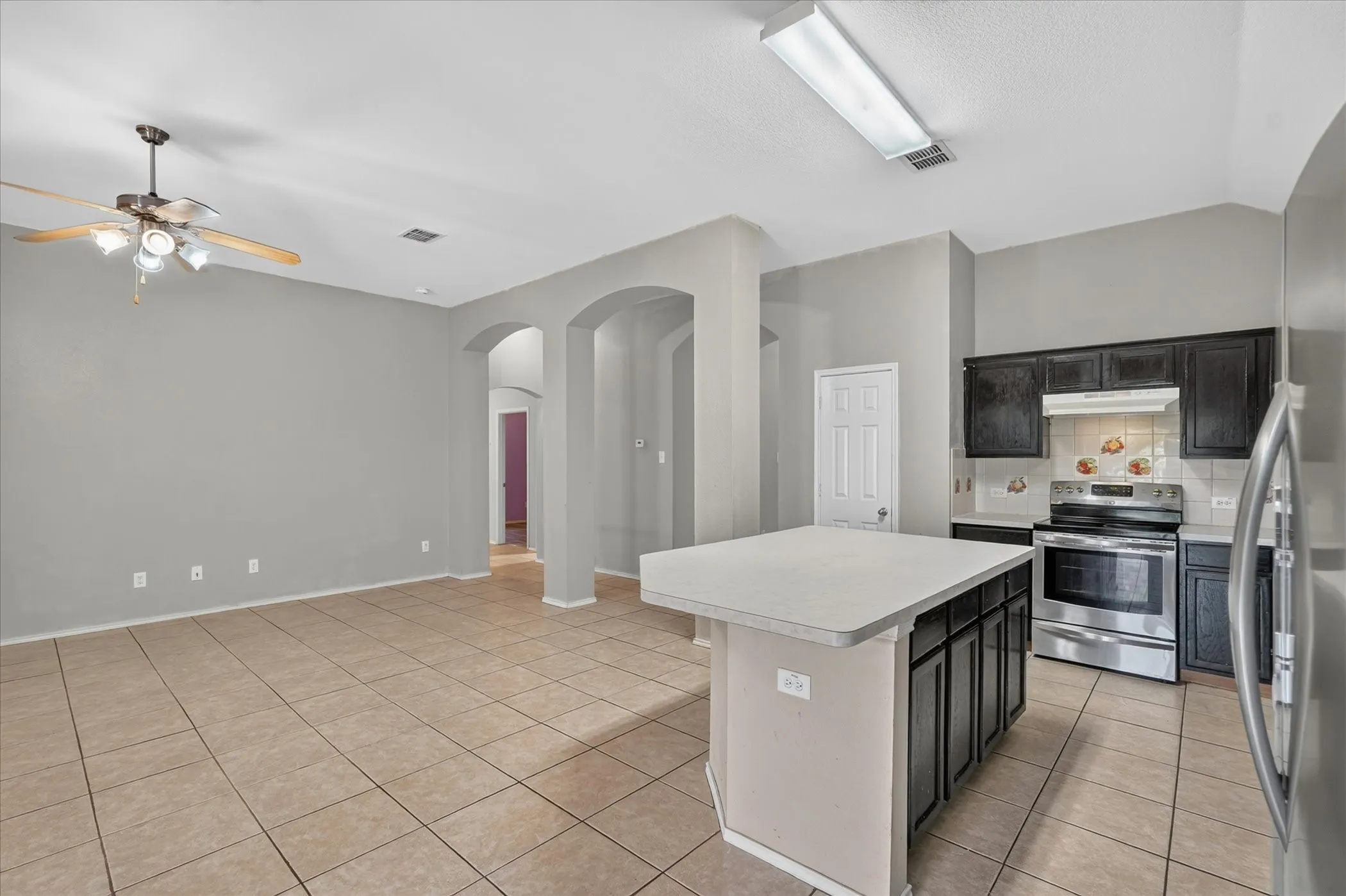 Kitchen and living area featuring a central island with a light-toned countertop, dark wood-finish cabinetry, stainless steel appliances, and tile flooring
