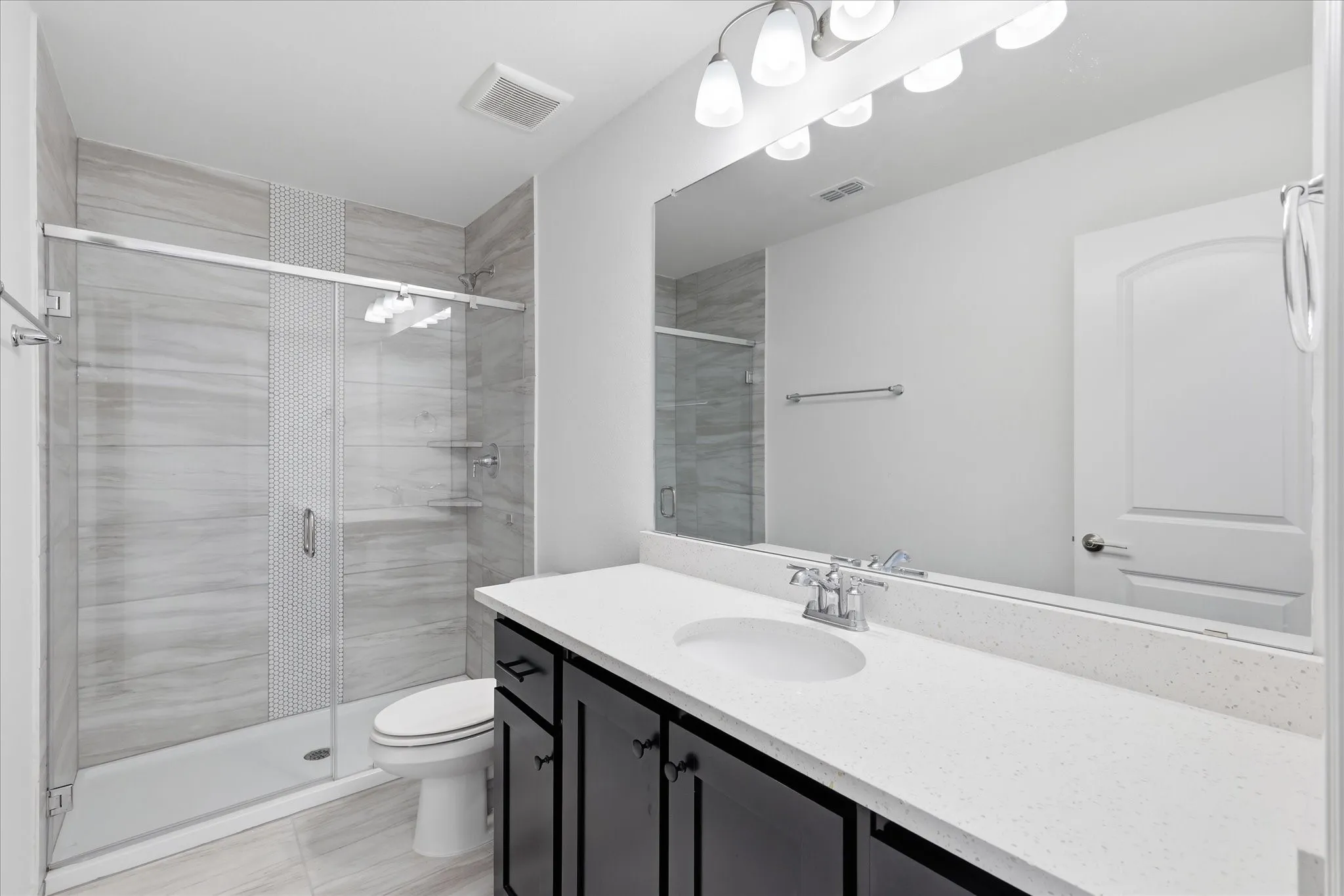 Spa-inspired primary bathroom featuring a frameless glass-enclosed walk-in shower with linear tile detailing, dual-sink vanity with dark cabinetry and white quartz countertop, chrome fixtures, LED vanity lighting, and polished tile flooring throughout