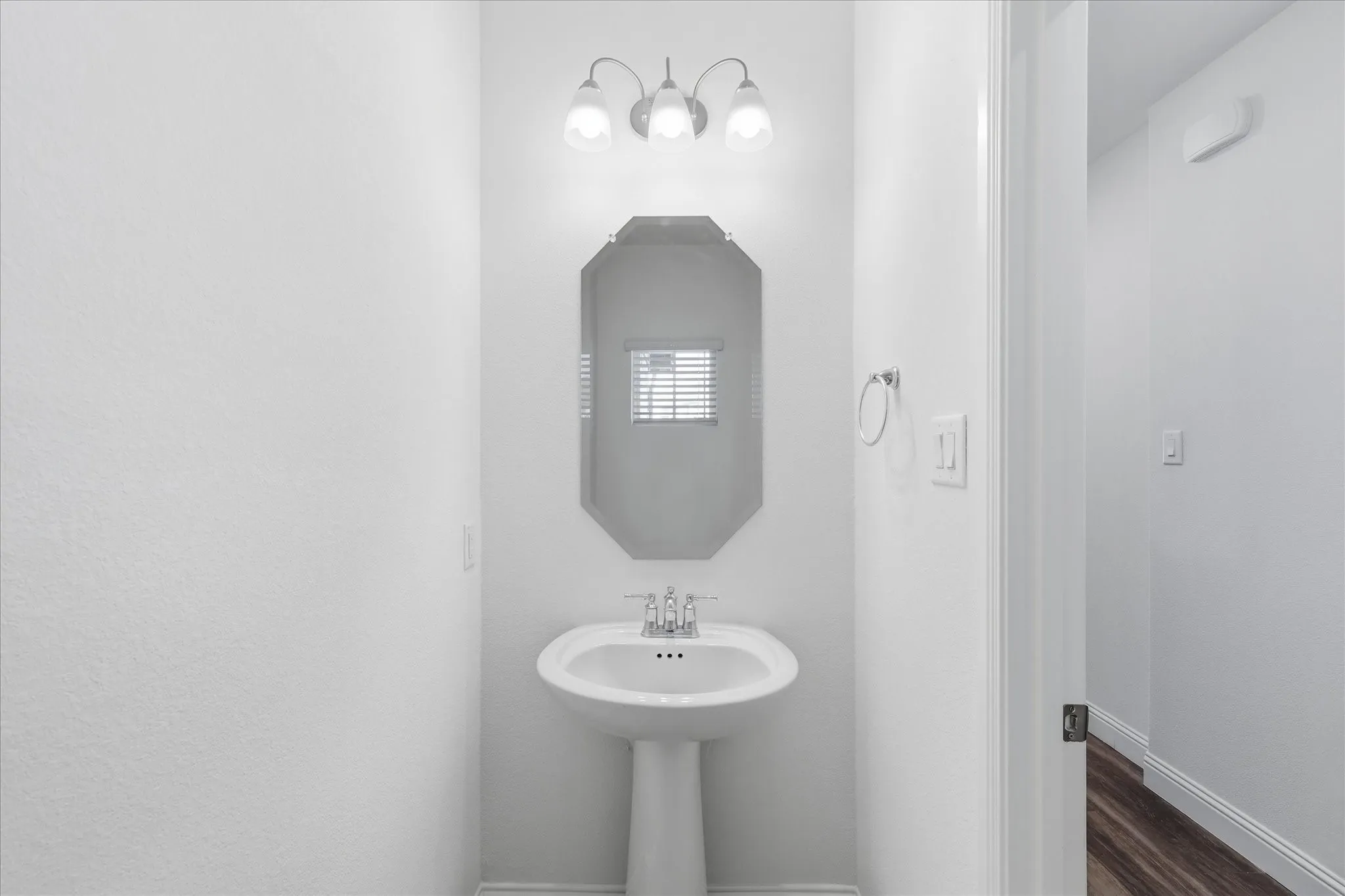 Elegant powder room featuring a pedestal sink with chrome fixtures, octagonal framed mirror with integrated lighting, crisp white walls, and wood-finish flooring