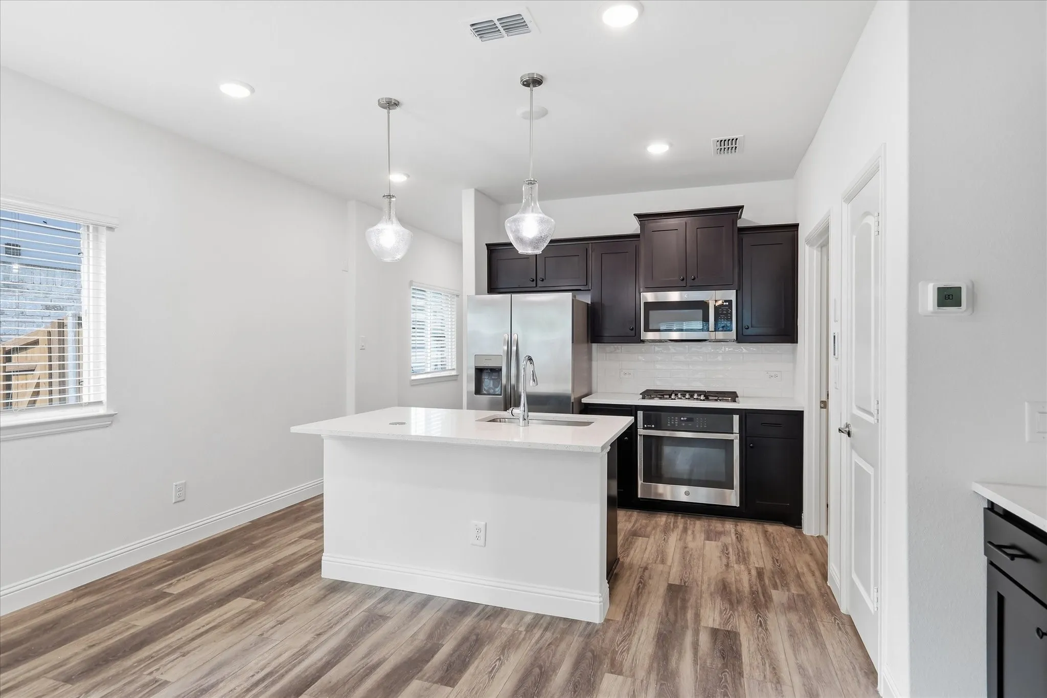Modern kitchen featuring dark wood cabinetry, stainless steel appliances, a white subway tile backsplash, and a central island with a white countertop and pendant lighting