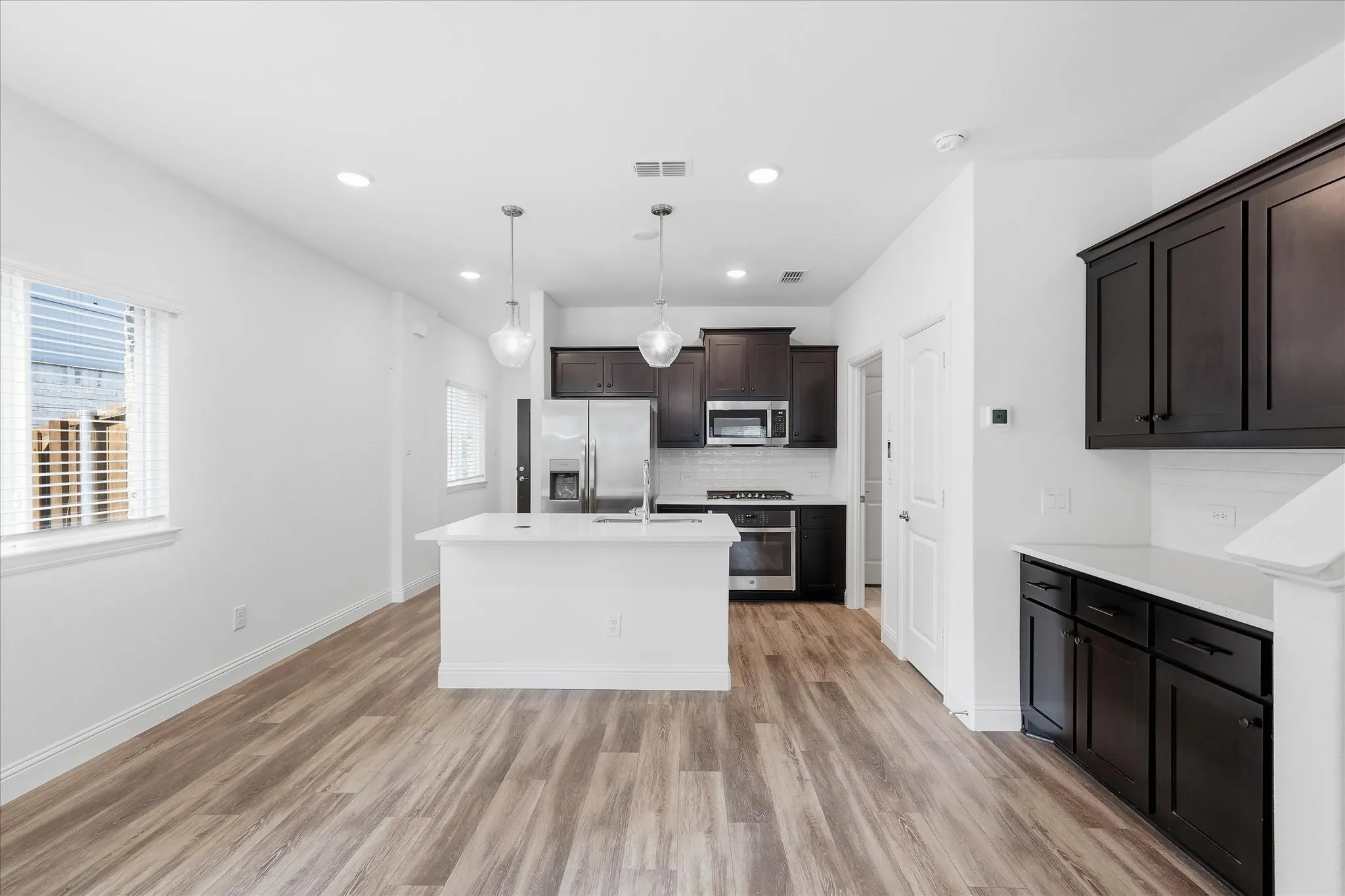 Modern open-concept kitchen and living area featuring a white quartz island with waterfall edge, stainless steel appliances including a built-in microwave, dark cabinetry with contrasting white perimeter counters, wood-finish plank flooring throughout, recessed ceiling lighting, and abundant natural light from multiple windows