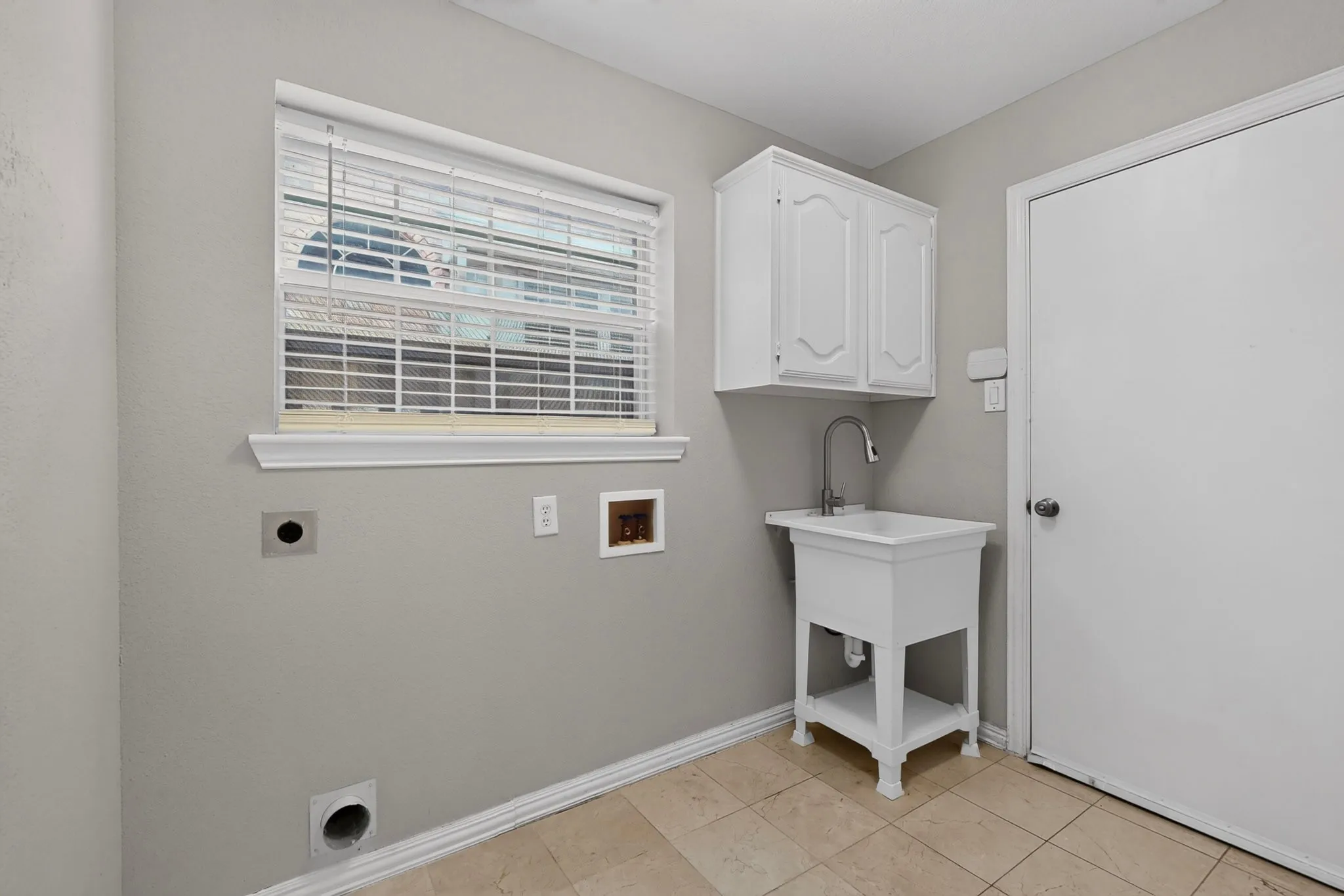 Utility room featuring a window with blinds, a white wash basin with a faucet, white upper cabinetry, and tile flooring