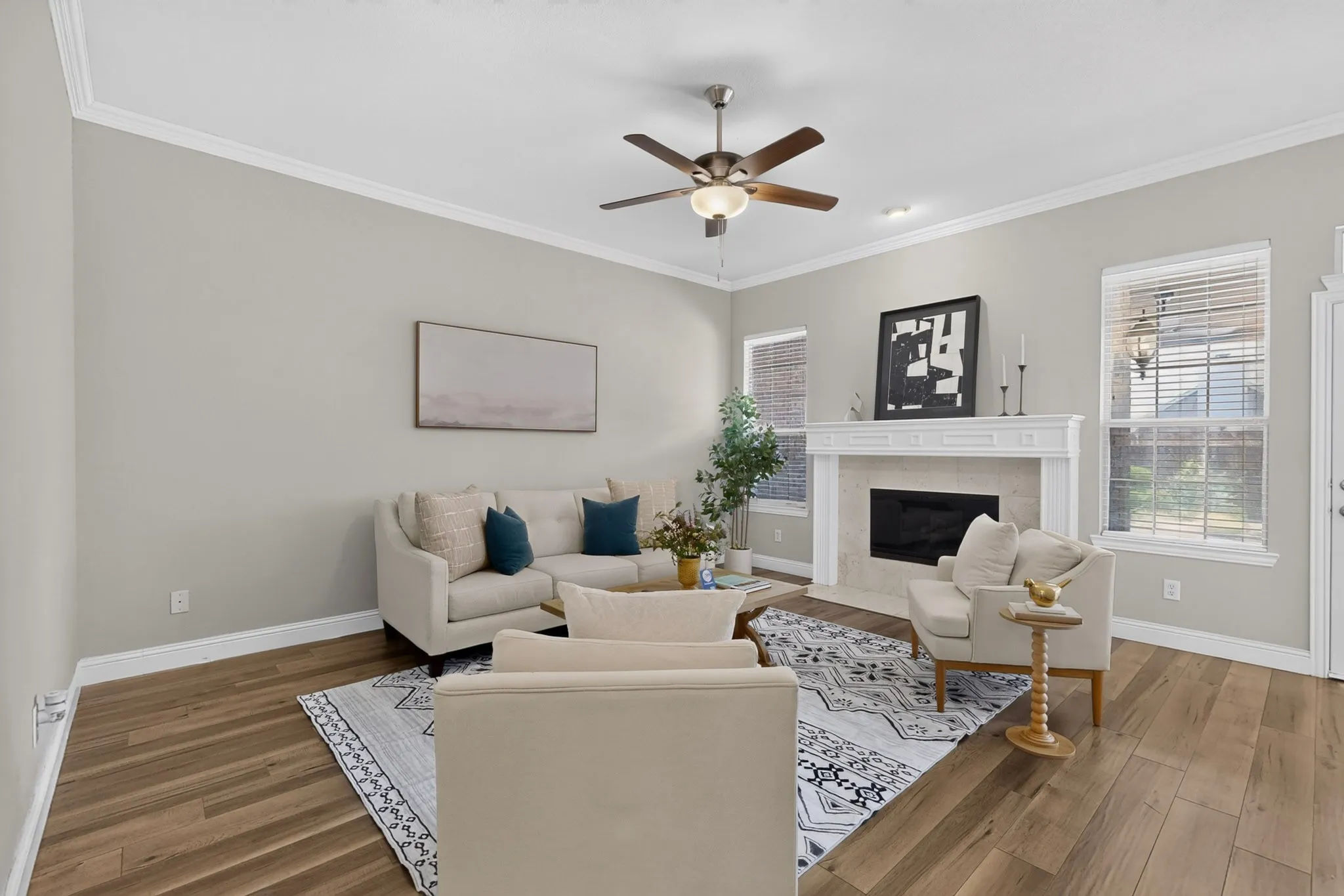 Spacious living area featuring light-toned walls, crown molding, wood-finish flooring, a decorative fireplace with a mantel, and a ceiling fan