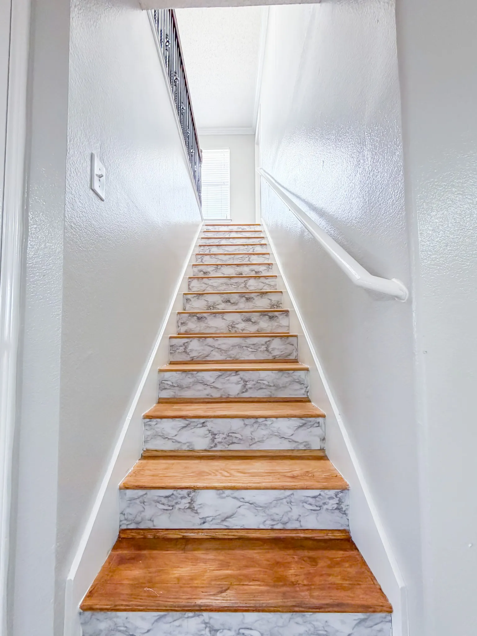 Staircase featuring wood treads and marble-look risers, complemented by a white handrail and a dark wrought iron banister
