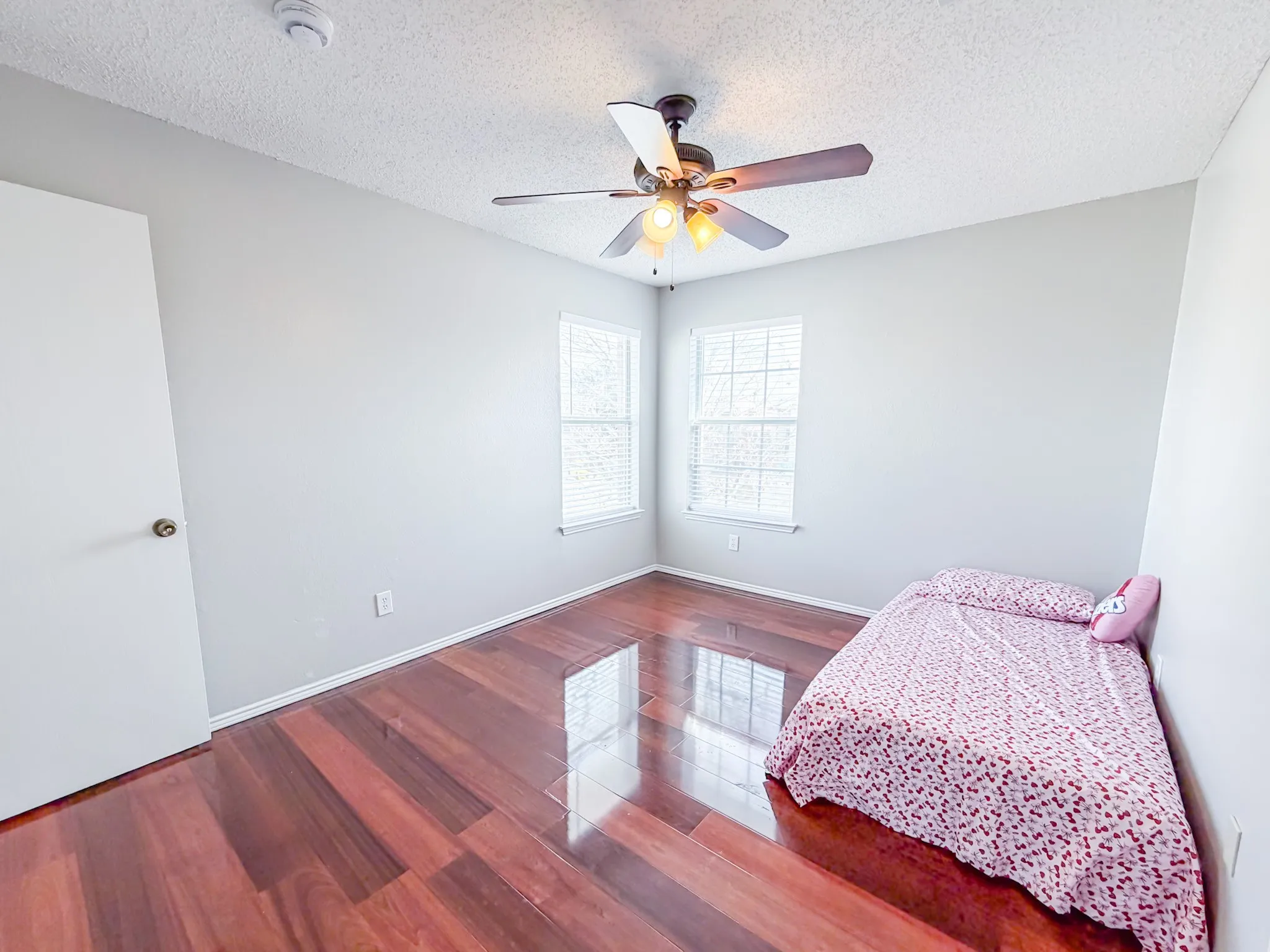 Room featuring polished wood-finish flooring, two large windows with blinds, and a ceiling fan with integrated lighting