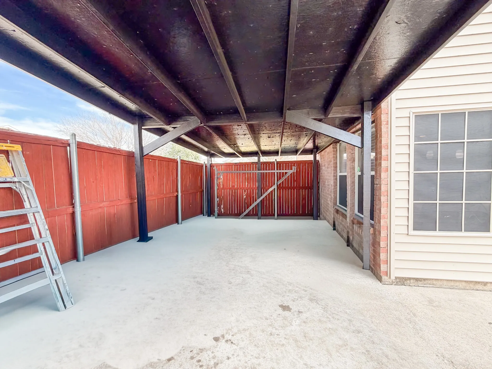 Covered outdoor space featuring a solid overhead structure, concrete flooring, and a red wood fence
