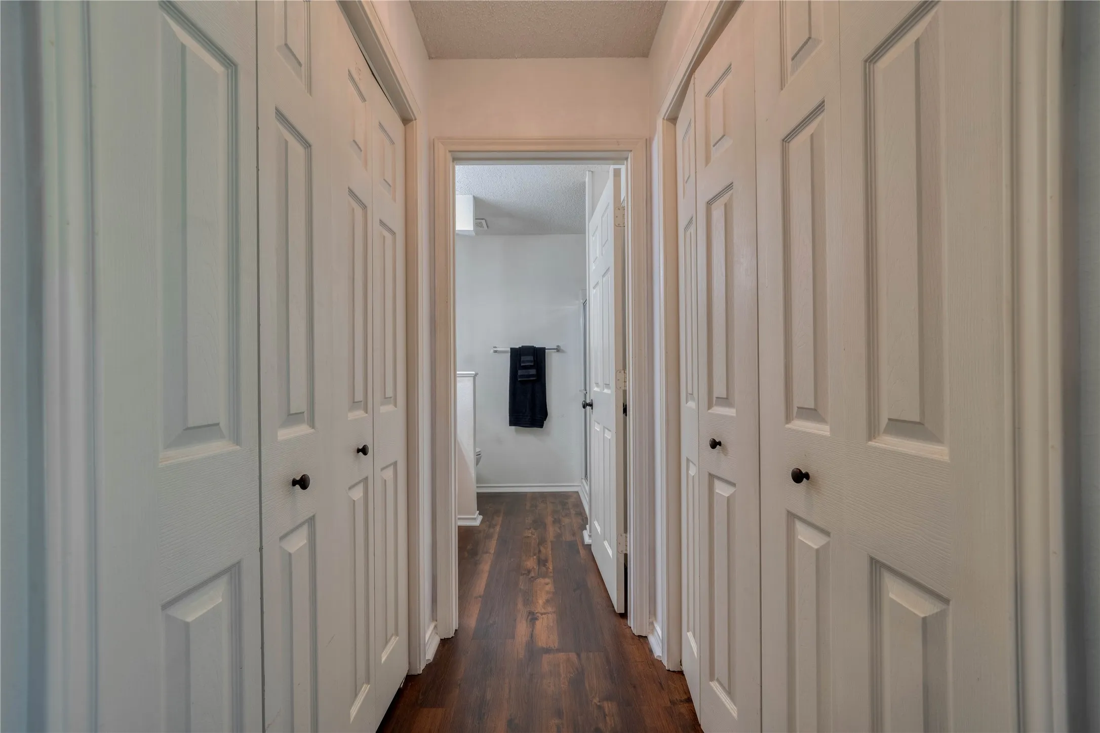 Hallway featuring wood-finish flooring, white paneled doors with black hardware, and an open doorway to another room