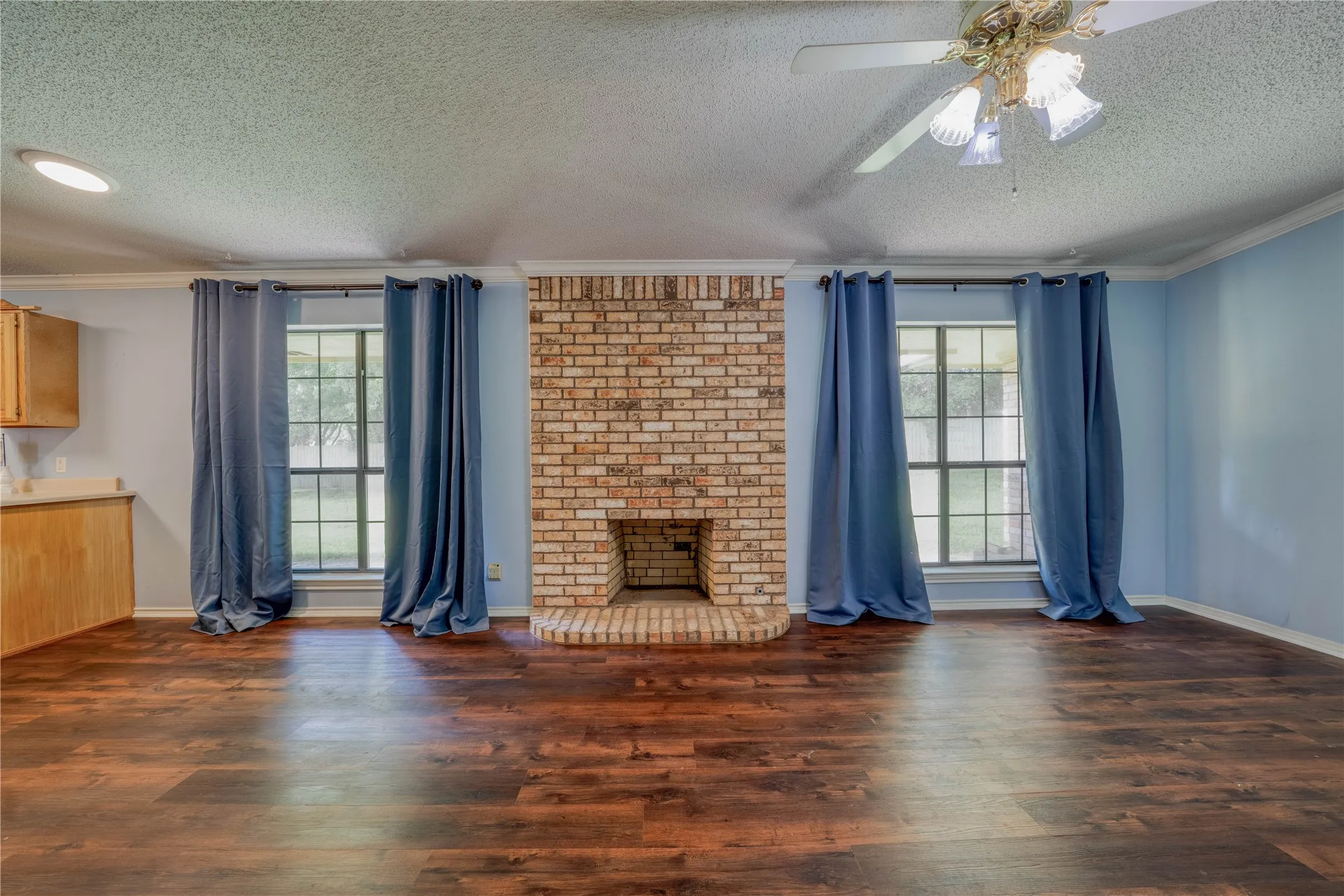 Spacious room featuring a prominent brick fireplace with a hearth, wood-finish flooring, and multiple windows with curtain rods