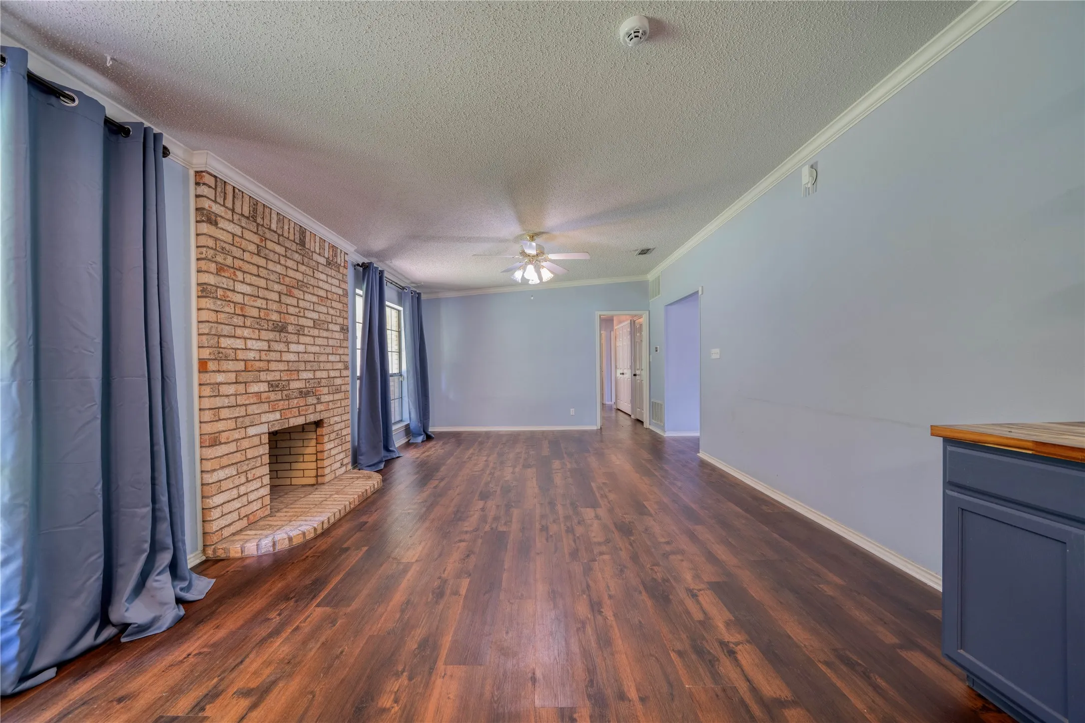 Spacious living area featuring wood-finish flooring, a brick fireplace with hearth, crown molding, and light blue walls