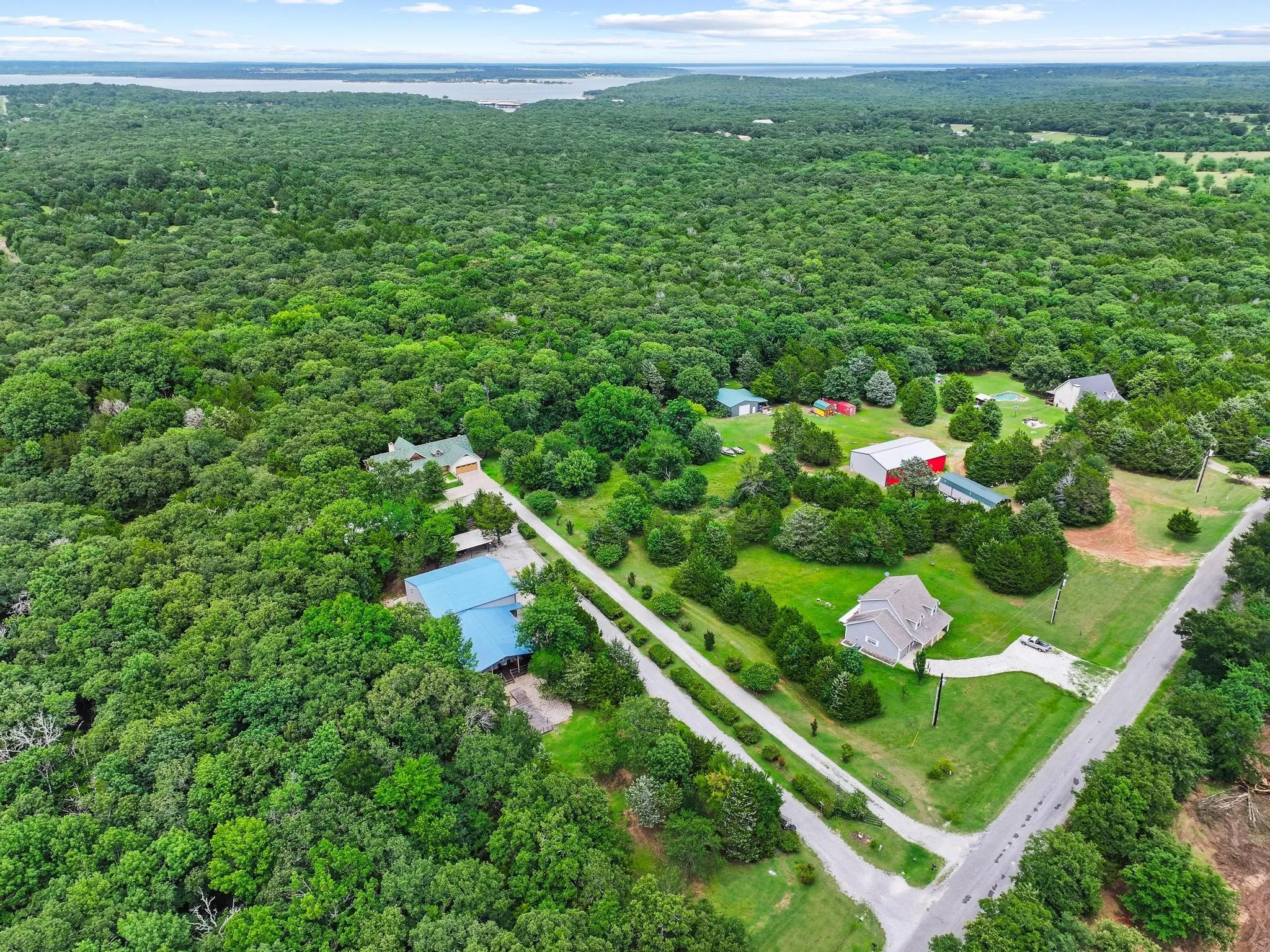 Expansive wooded acreage featuring a distant body of water, multiple residential structures with distinct roofing, a red barn, and established driveways