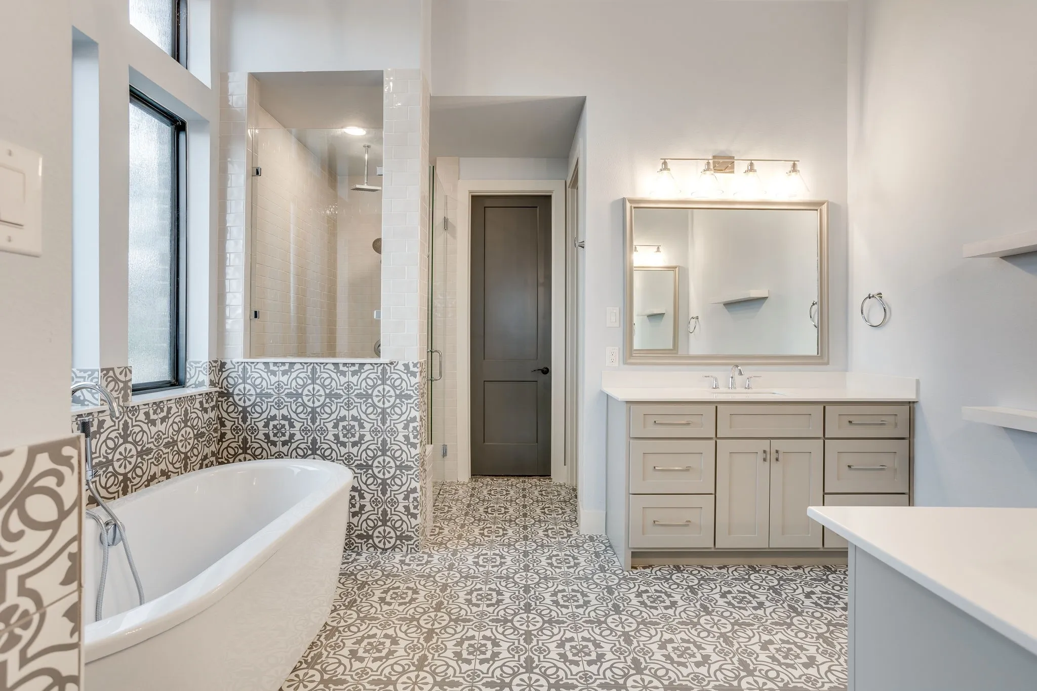 Bathroom featuring a freestanding soaking tub, patterned floor and wall tile, a glass-enclosed shower, a double vanity with a light-colored countertop, and a framed mirror with overhead lighting
